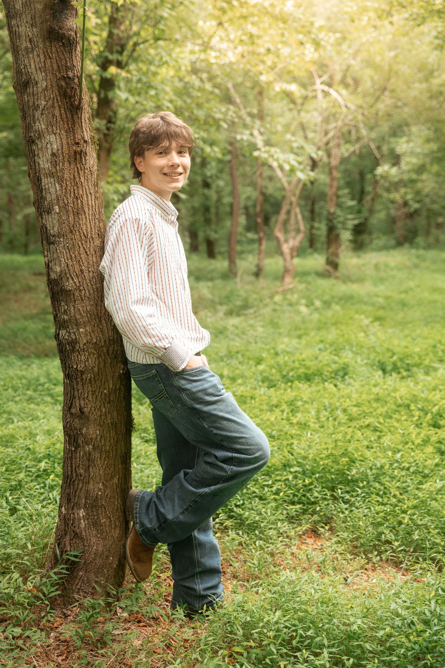 Smiling teenage boy leaning against a tree trunk in a lush green forest wearing jeans and a striped shirt.
