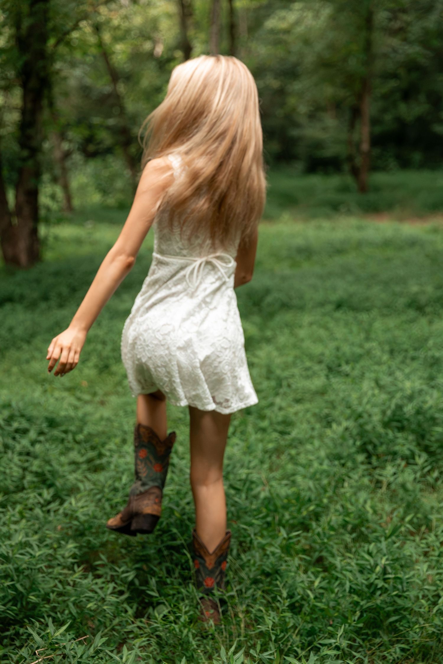 Young girl in white lace dress and cowboy boots walking through lush green field toward forest.