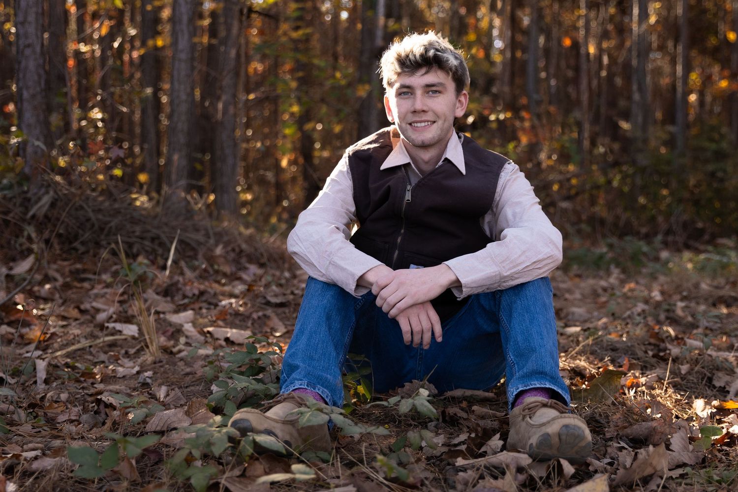 Young man in vest and jeans sitting on autumn leaves in a forest, smiling for a portrait photo- Senior portraits Northwest GA
