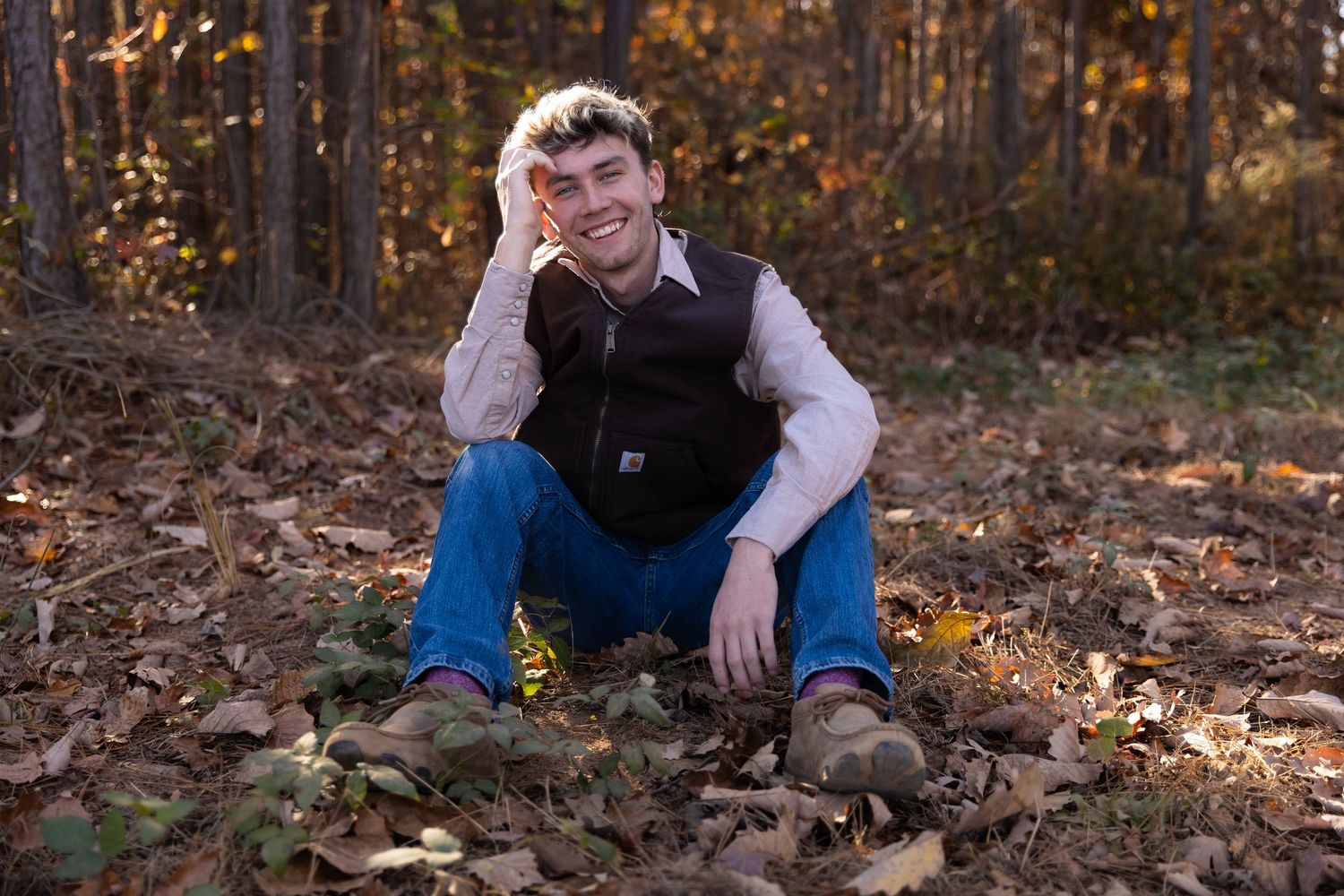 Young man smiling, sitting on autumn leaves in a forest wearing jeans and a vest for a senior portrait Cartersville GA Senior portraits