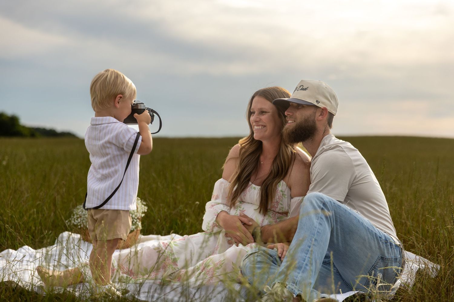 A young family enjoys a picnic in a green field as their toddler son photographs them with a camera.
