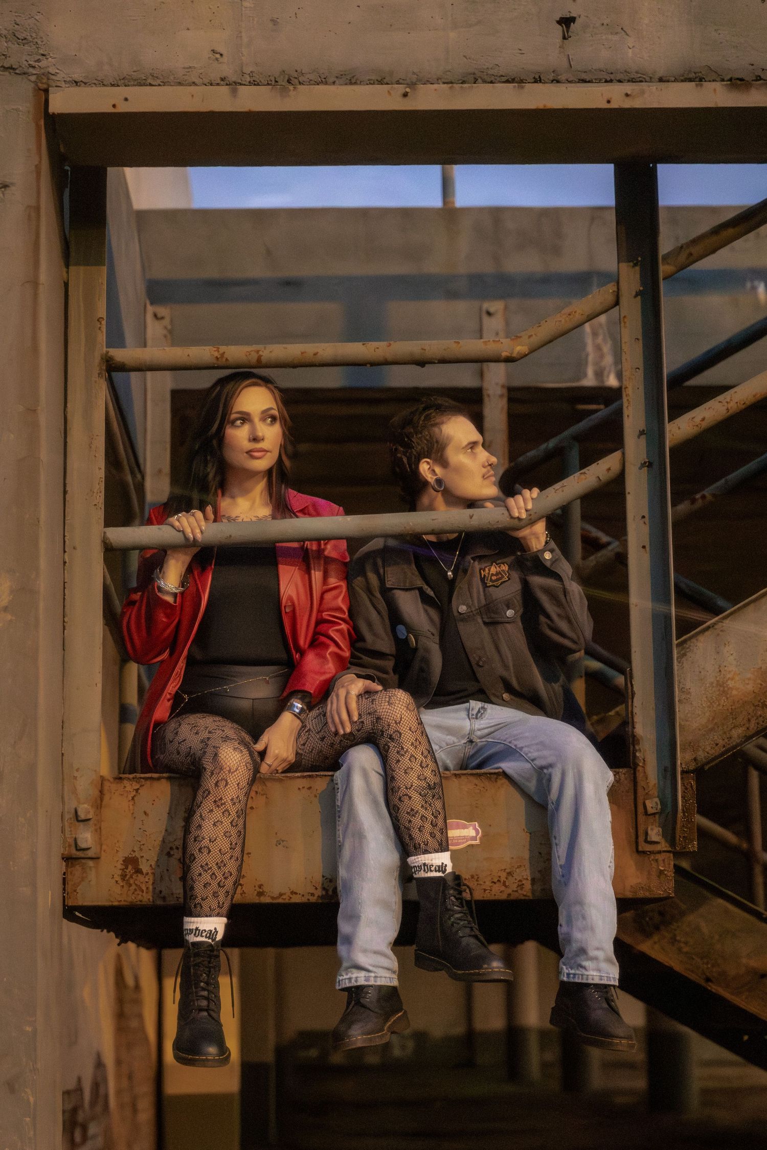A young couple sits on rustic metal stairs in an industrial setting, wearing edgy fashion including a red jacket and fishnet tights.
