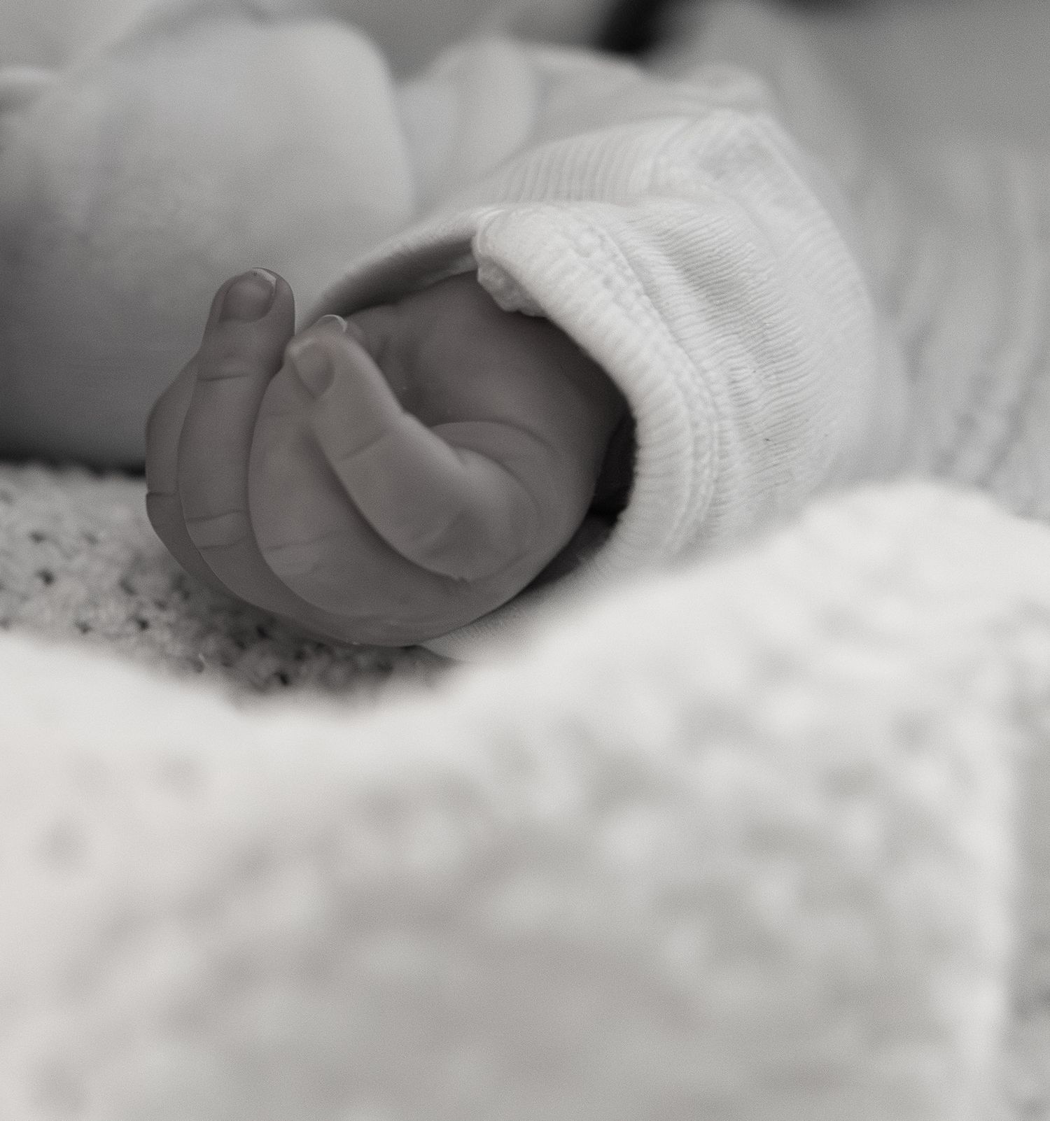 Close-up black and white photo of a newborn baby's tiny hand in a white knit sleeve on a soft blanket.