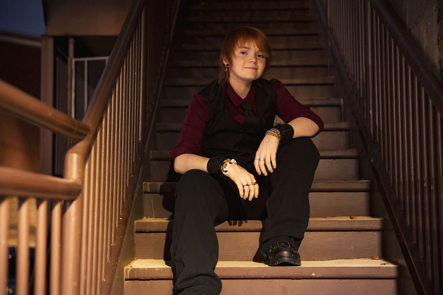 Young person sitting relaxed on outdoor stairs at night, wearing black pants and dark red top with gold bracelets. Rome, GA