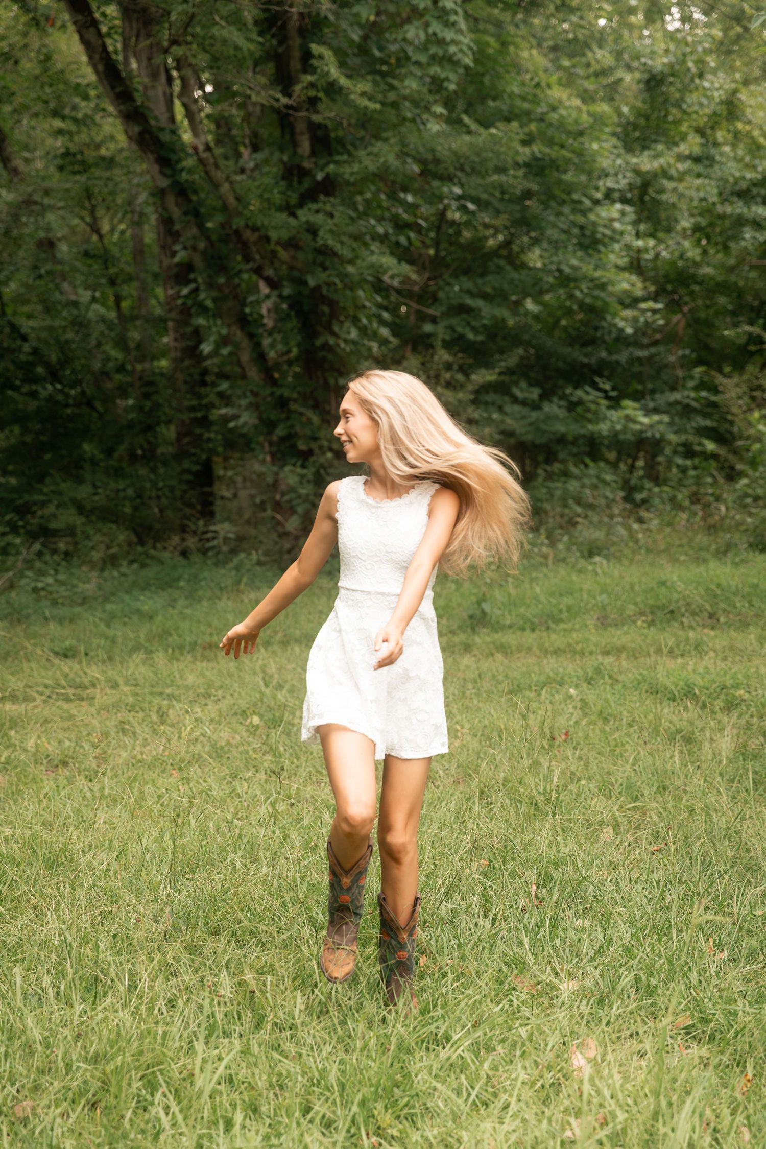Carefree blonde girl in white dress and cowboy boots twirling in green meadow with hair flowing.