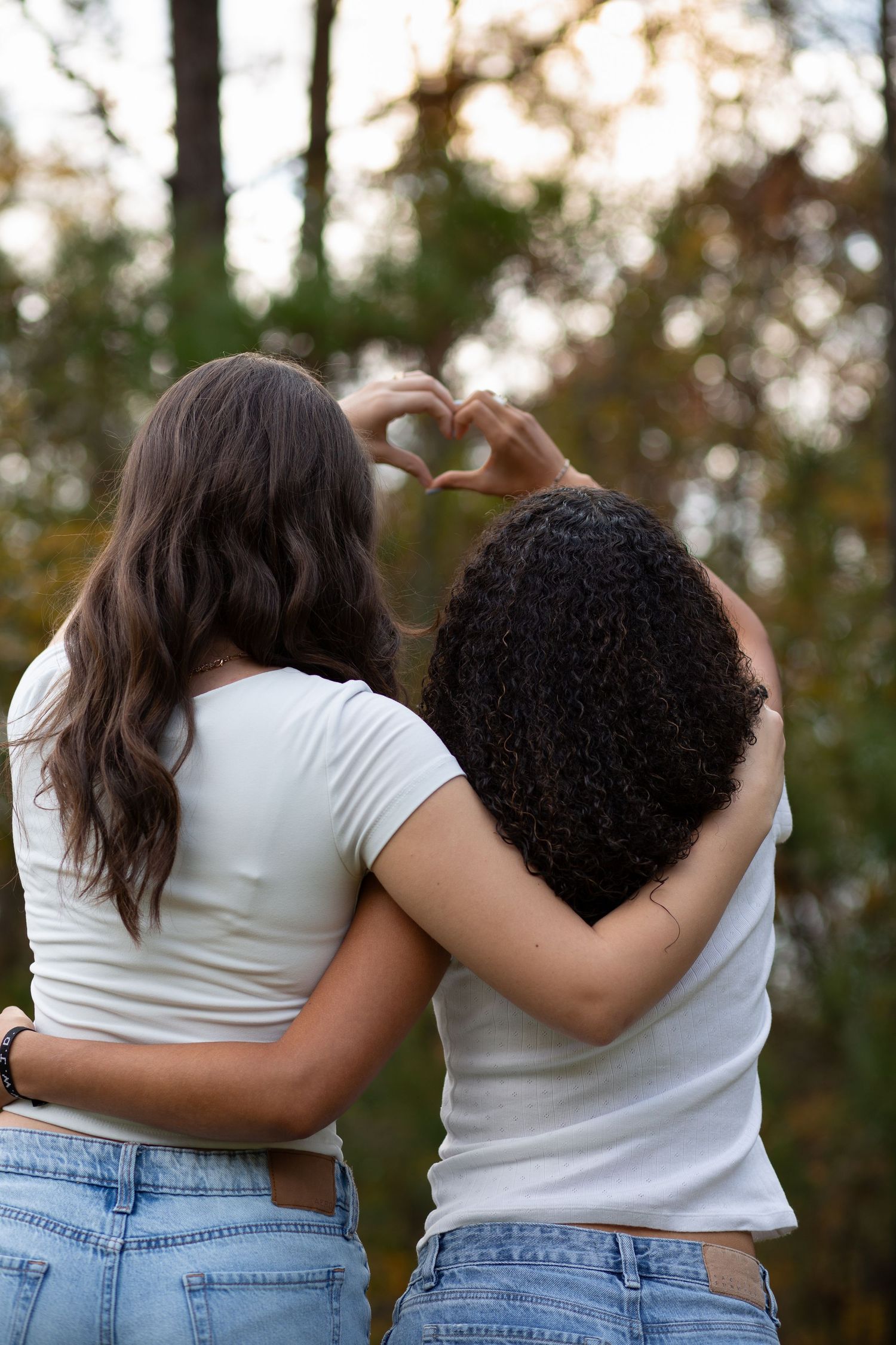 Two women in white tees and jeans embrace from behind, forming a heart shape with their hands outdoors.