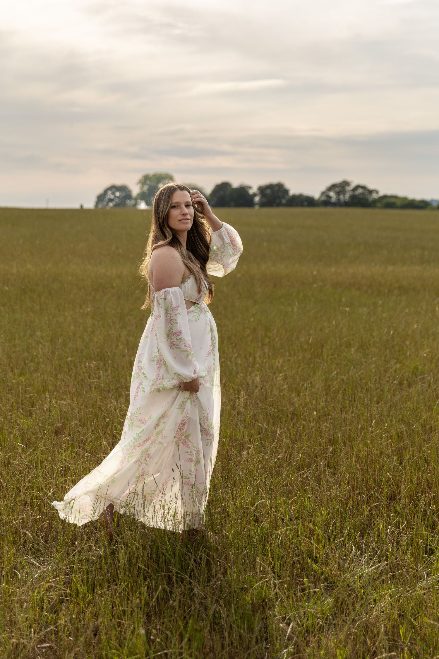 A woman in a flowing floral white off-shoulder dress stands gracefully in a green field under a cloudy sky.