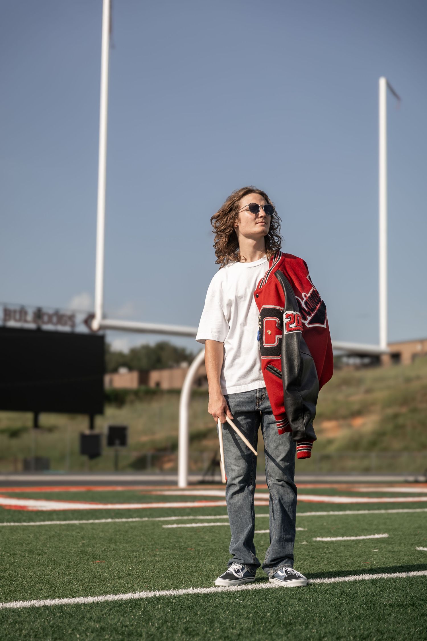 Teen boy with curly hair holds drumsticks on football field wearing varsity jacket and sunglasses. Cedartown High School, Cedartown GA