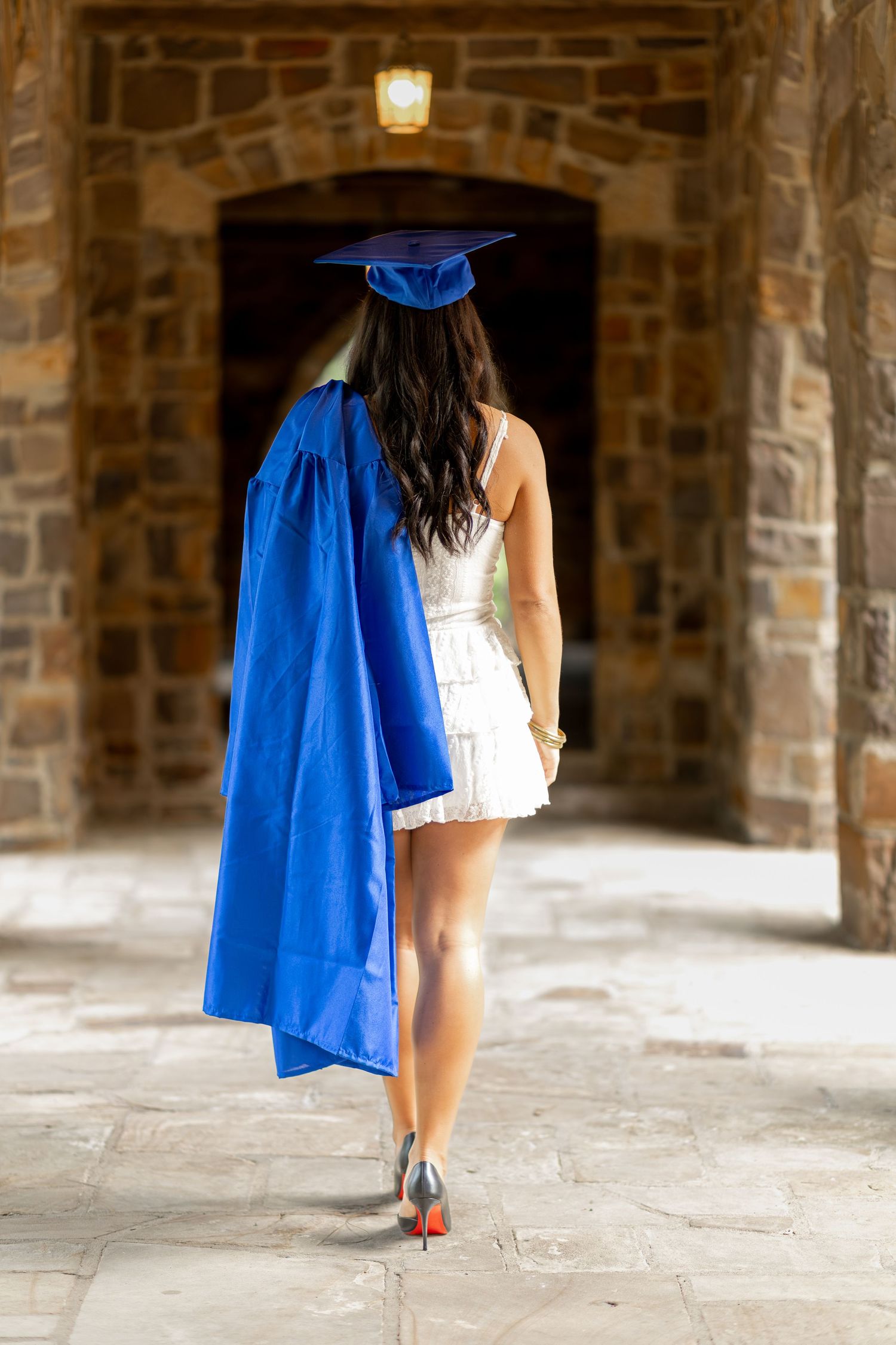 Graduate woman in blue cap and gown walking toward stone archway entrance in white dress and heels. Senior Photographer Rome, GA