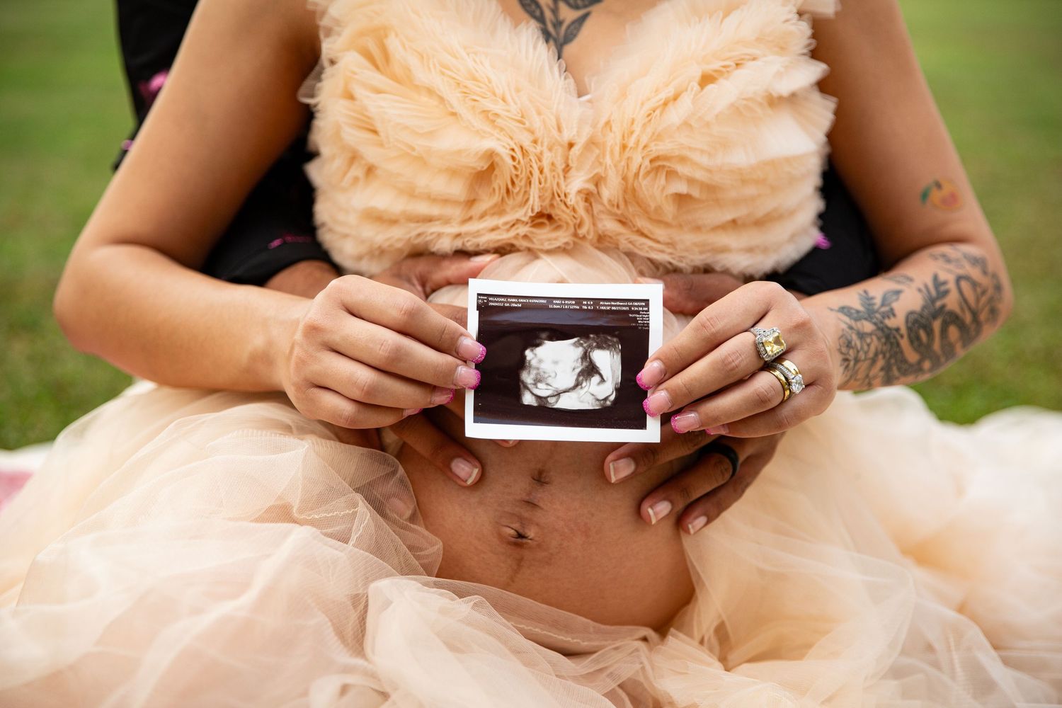 Pregnant woman in peach tulle dress holds ultrasound photo over baby bump, showing tattooed arms and ring. Berry College Rome GA