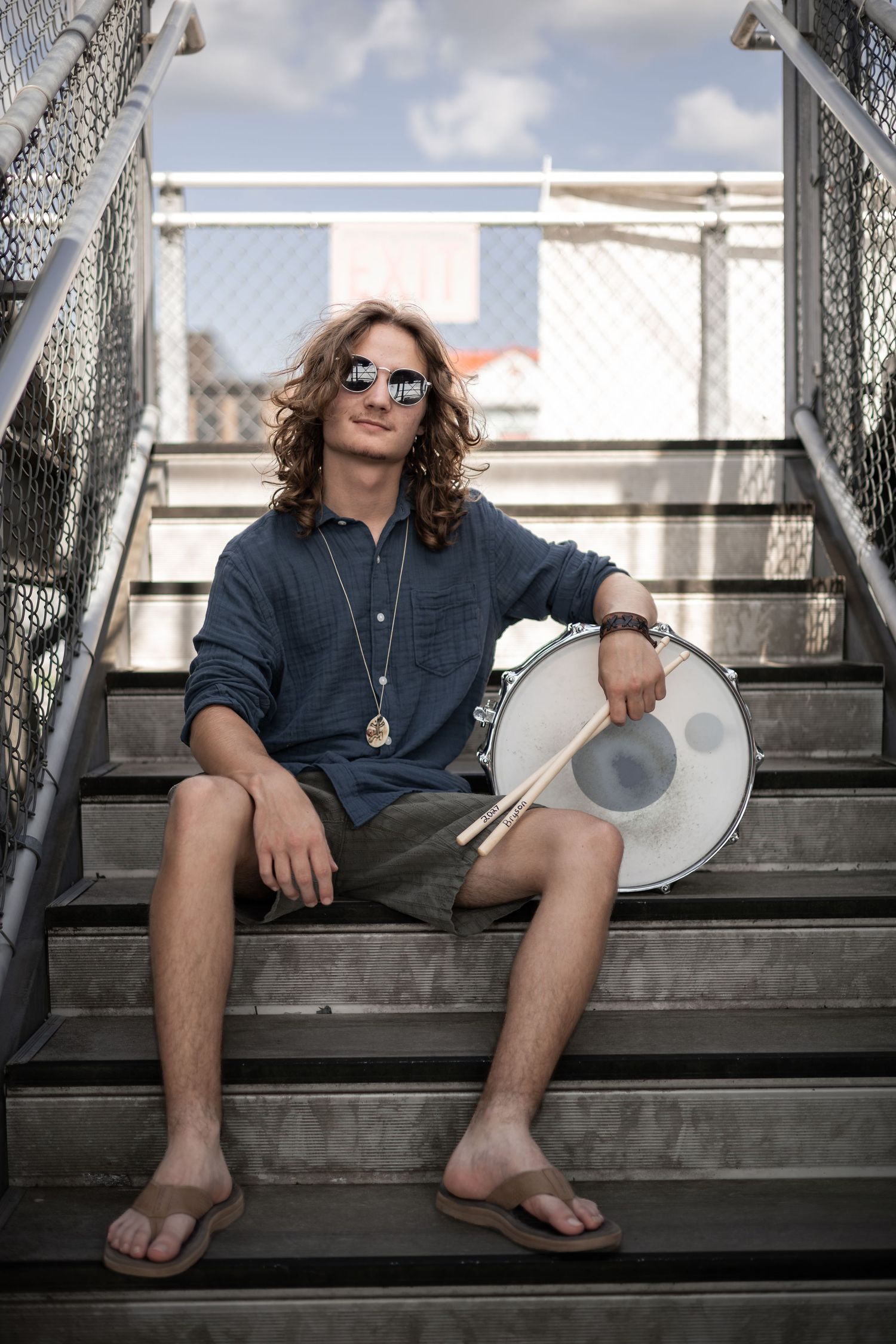 Young male drummer with long hair and sunglasses sitting on outdoor stairs holding drumsticks and a snare drum.  Senior Portraits Cedartown