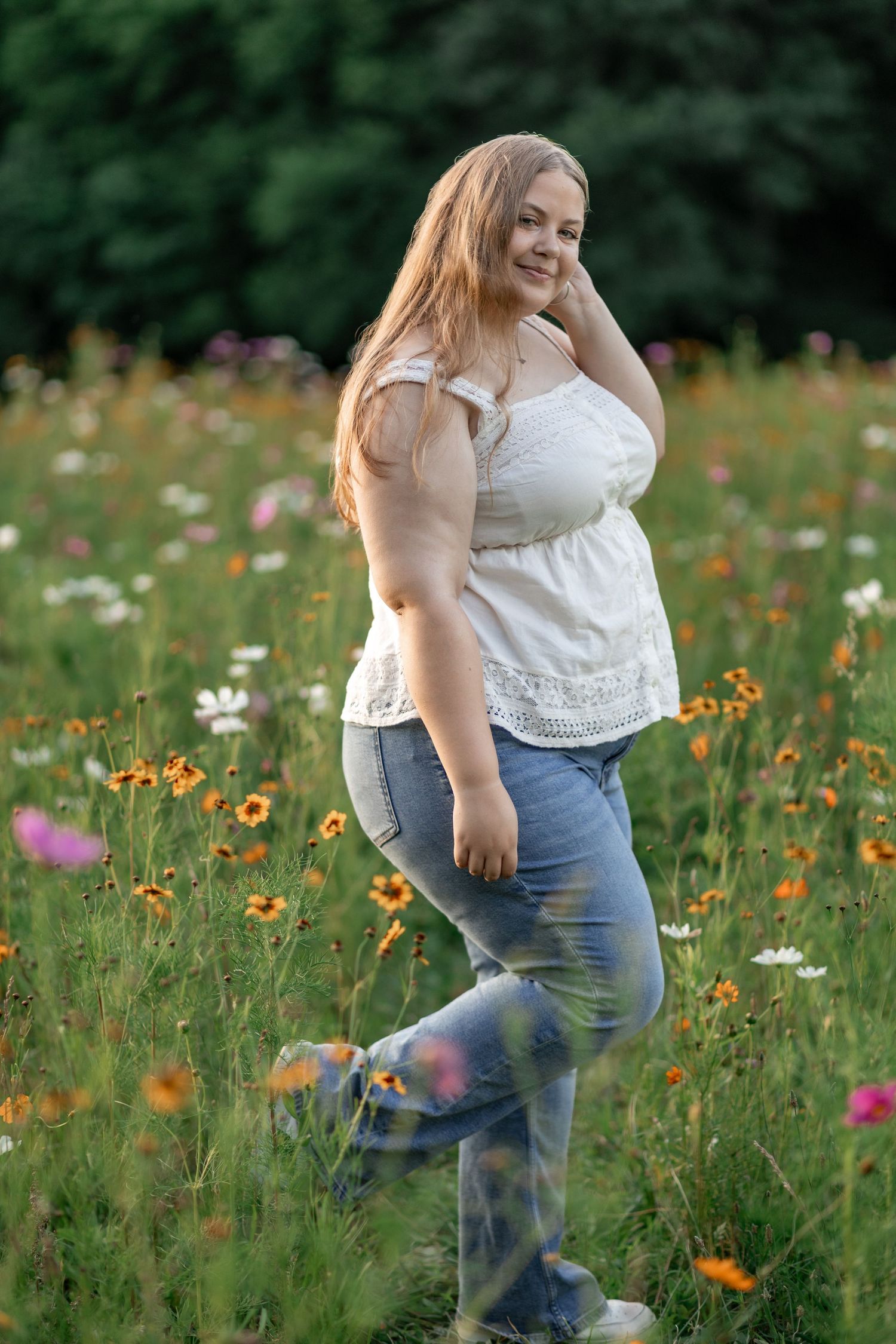 A smiling plus-size woman in white top and jeans poses in a vibrant wildflower meadow with colorful blooms.