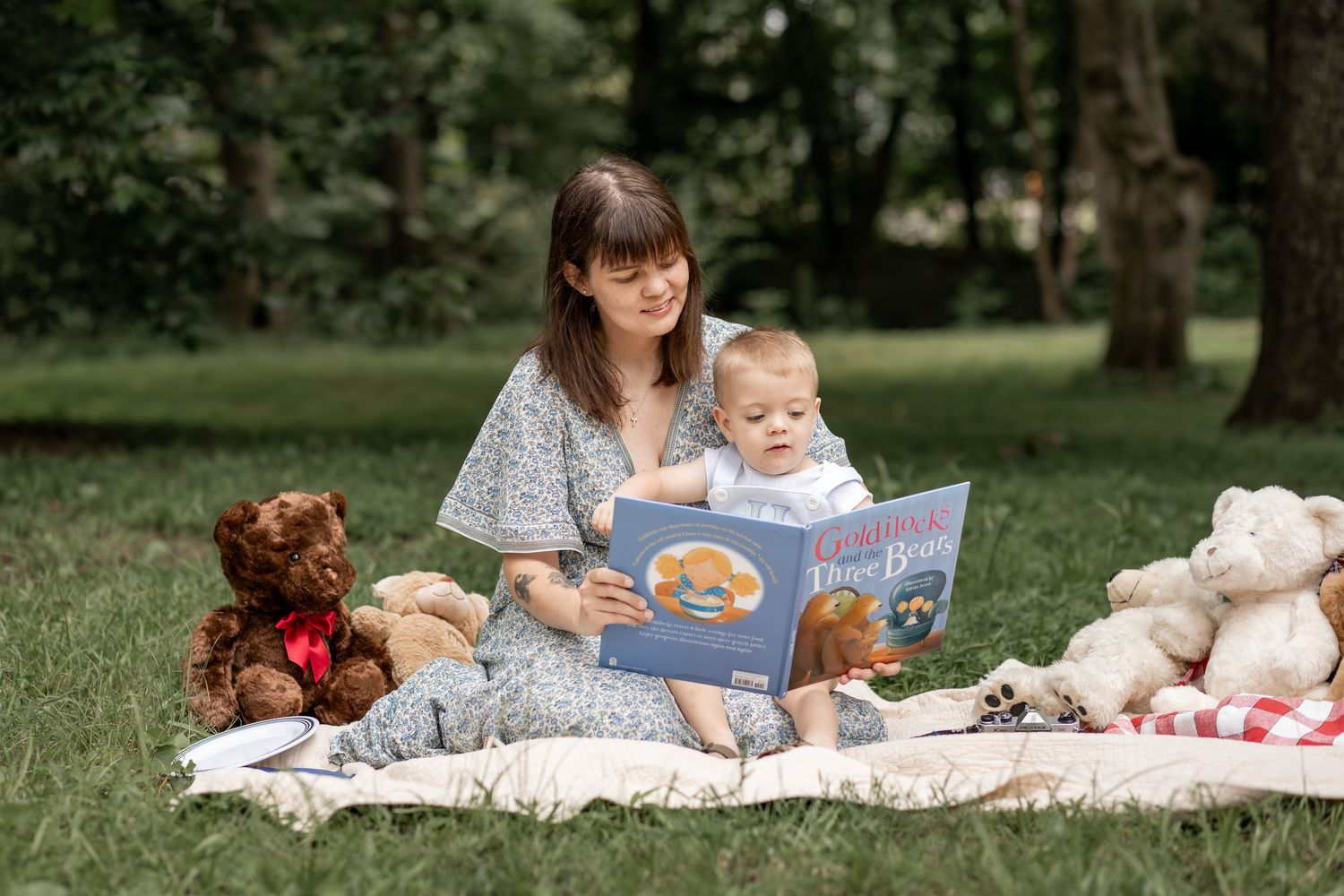 Mother reads Goldilocks and the Three Bears to baby on picnic blanket surrounded by teddy bears in a park.