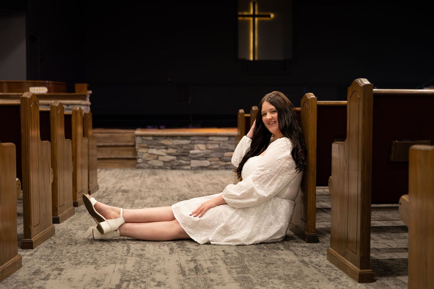Young woman in white dress sitting between church pews, smiling, with a wooden cross on the wall behind her Cartersville, GA