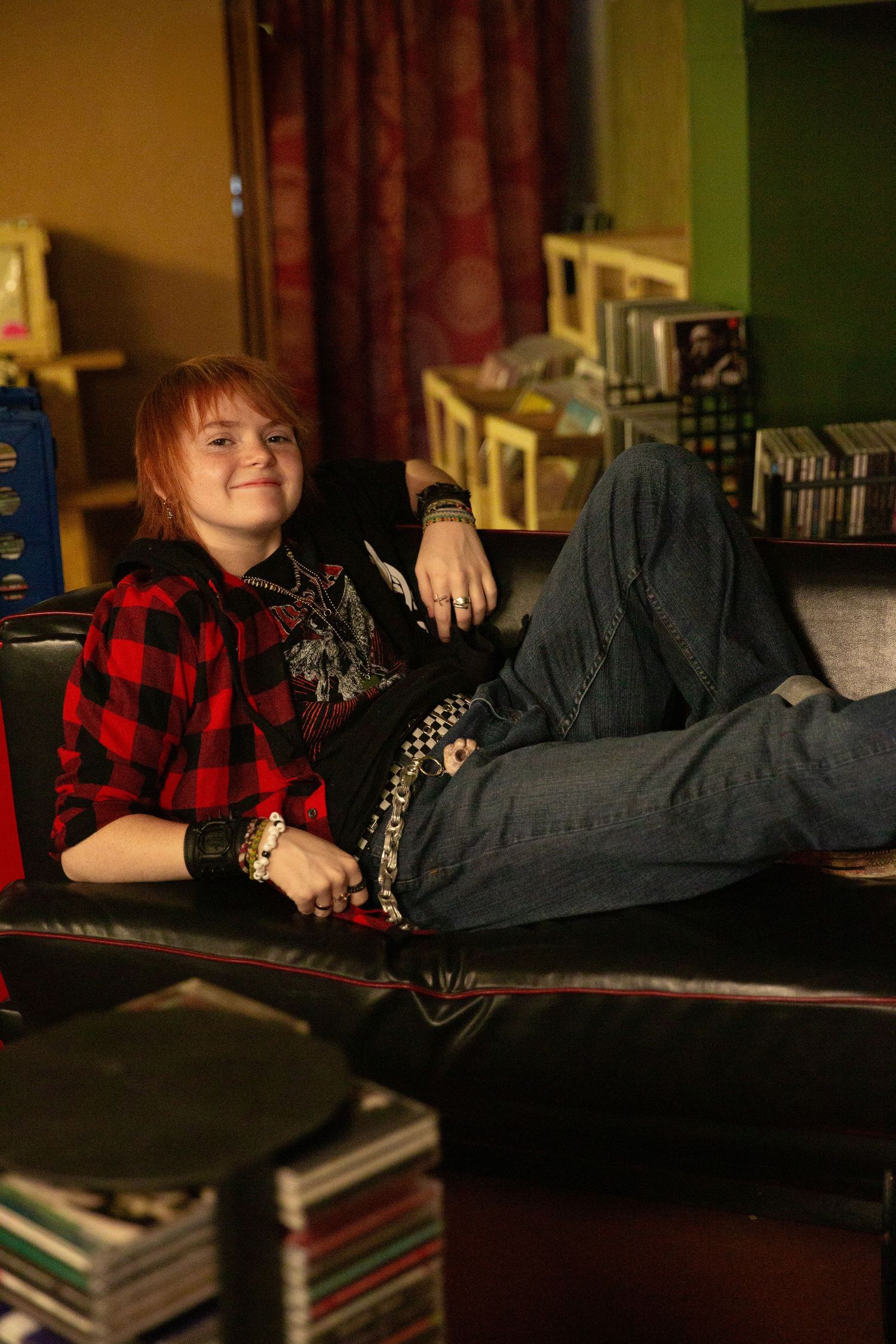 Young person with red hair relaxing on a black leather sofa, wearing a plaid shirt and jeans in a cozy room. Downtown record store Rome, GA