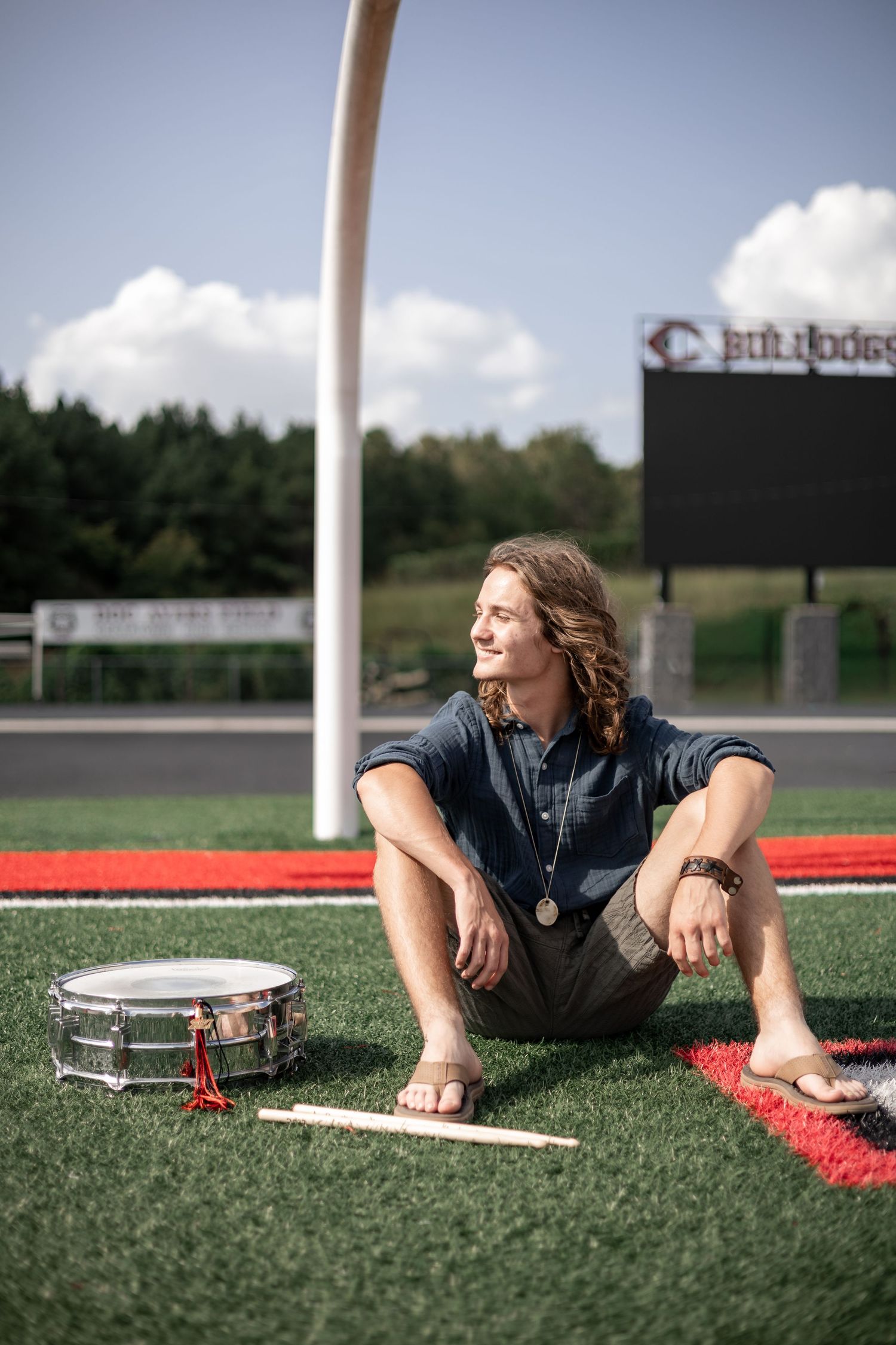 Long-haired teen sits on football field turf with snare drum and drumsticks, Bulldogs scoreboard visible behind him. Cedartown High SchoolGA