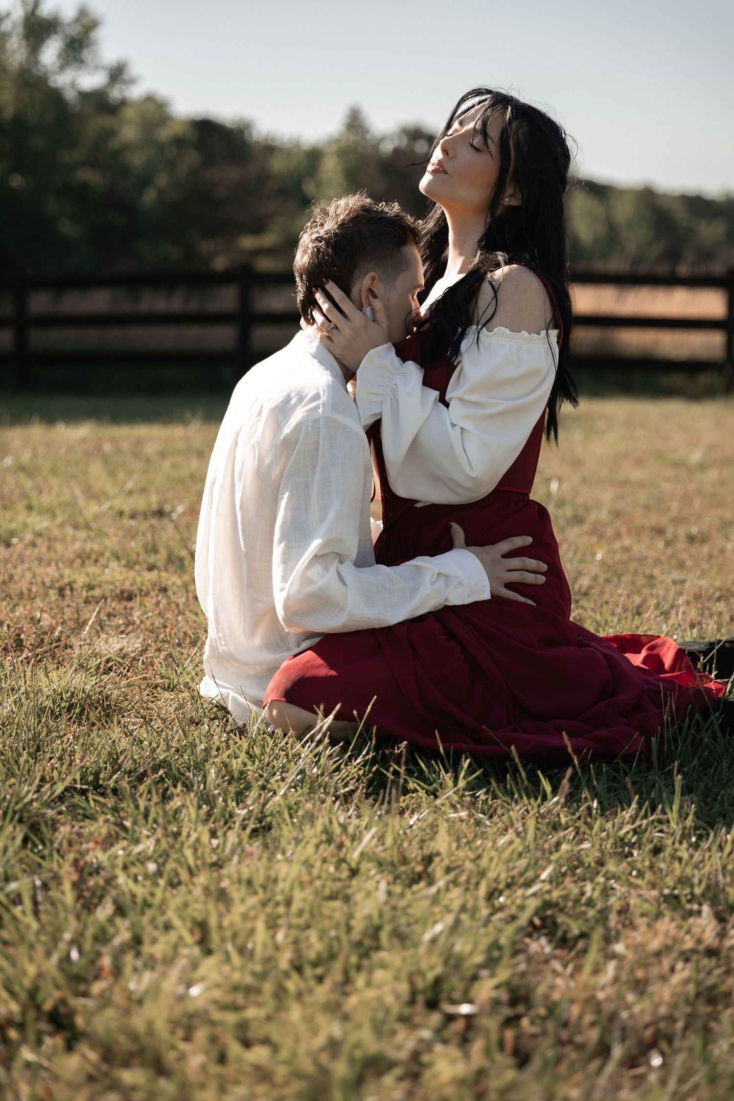 Romantic couple sitting on grass field, woman in red dress embracing man in white shirt during golden hour outdoors. Berry College Rome, GA