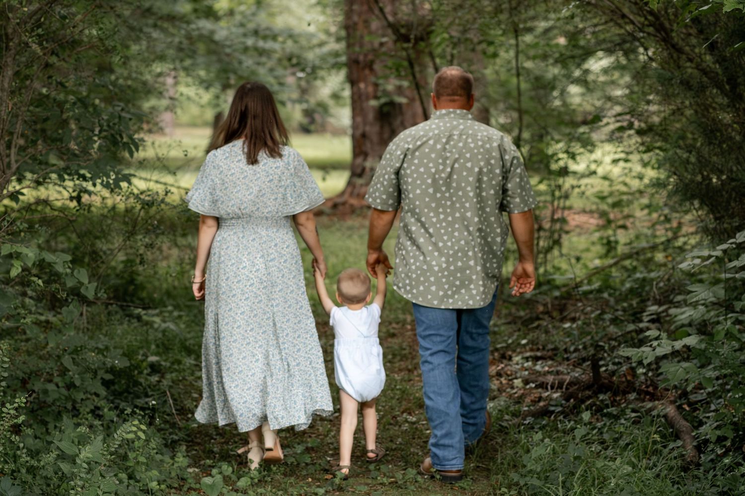 Family of three walking hand-in-hand through a lush green forest path, parents in floral outfits with toddler between them.