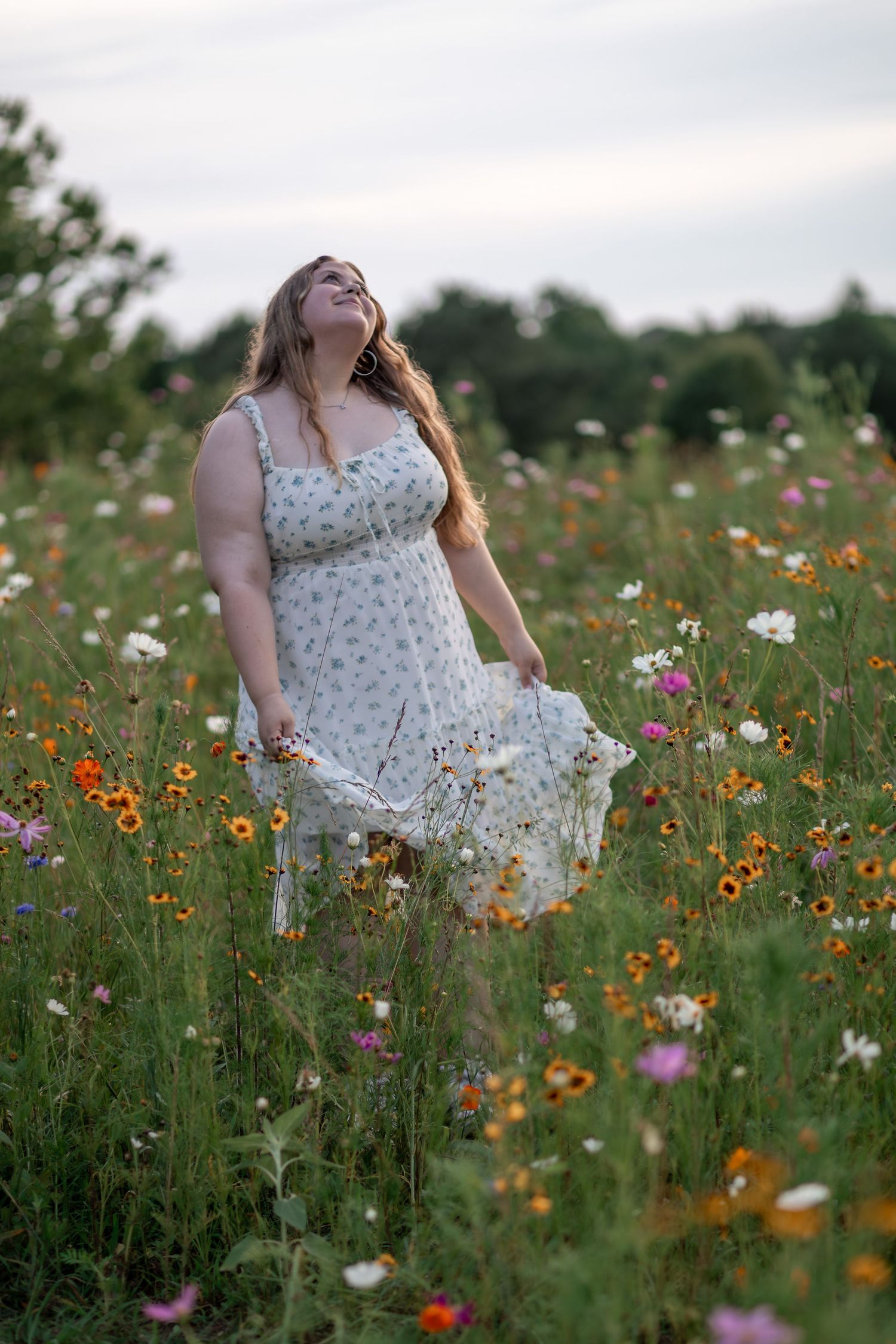 Woman in floral dress standing in colorful wildflower field, head tilted back, long hair flowing in breeze.