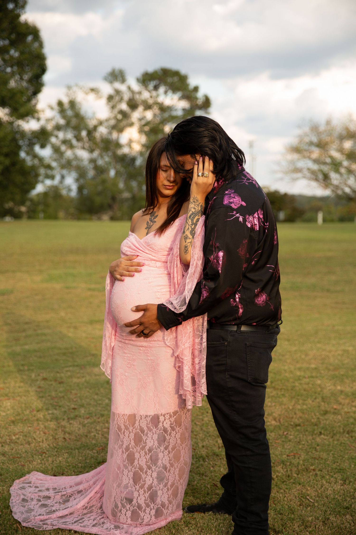 Couple sharing tender moment outdoors, pregnant woman in pink lace gown, partner in floral jacket, green park setting. Rome GA