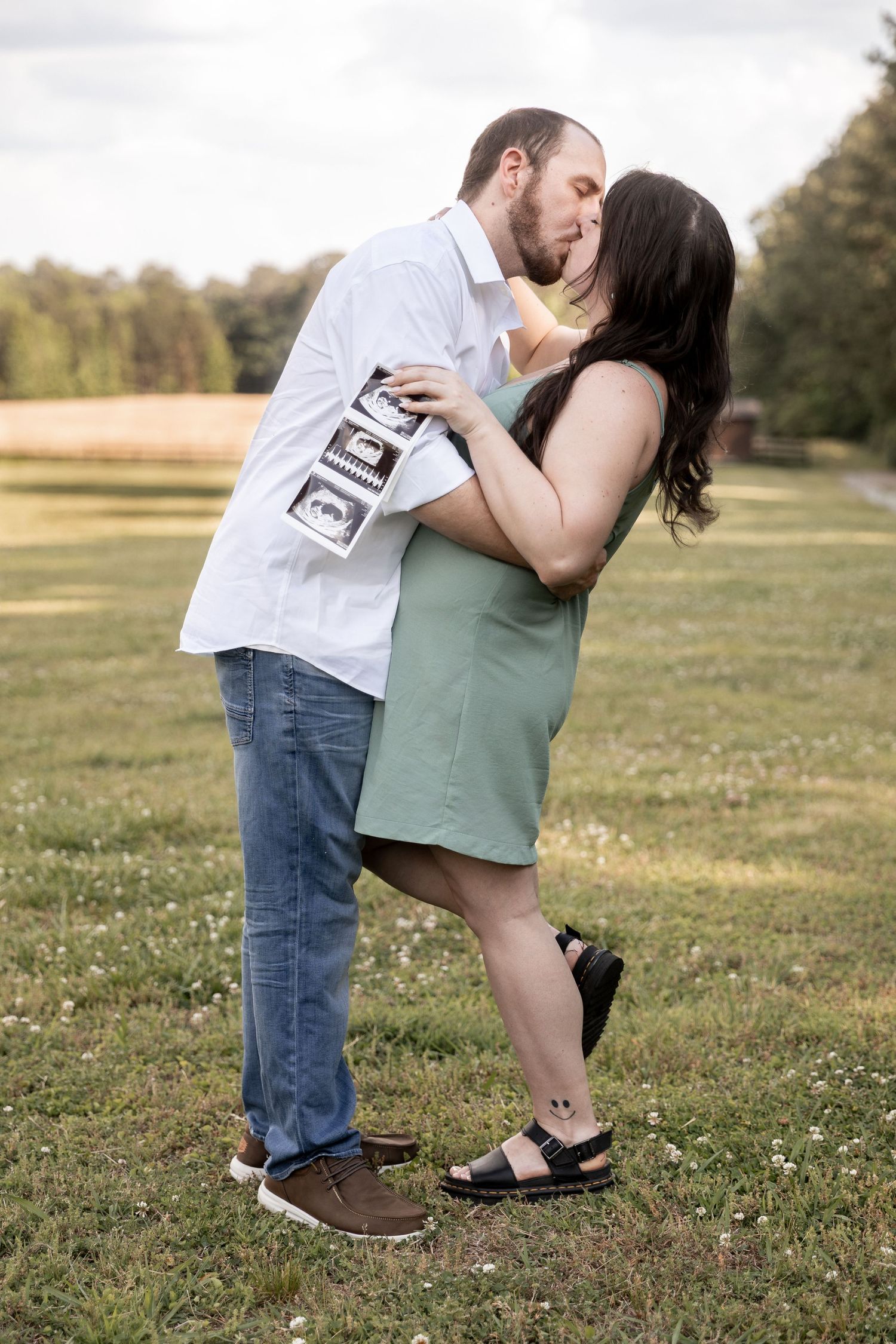 Couple kissing outdoors in a field, holding ultrasound photos, announcing pregnancy in a romantic maternity photo shoot.