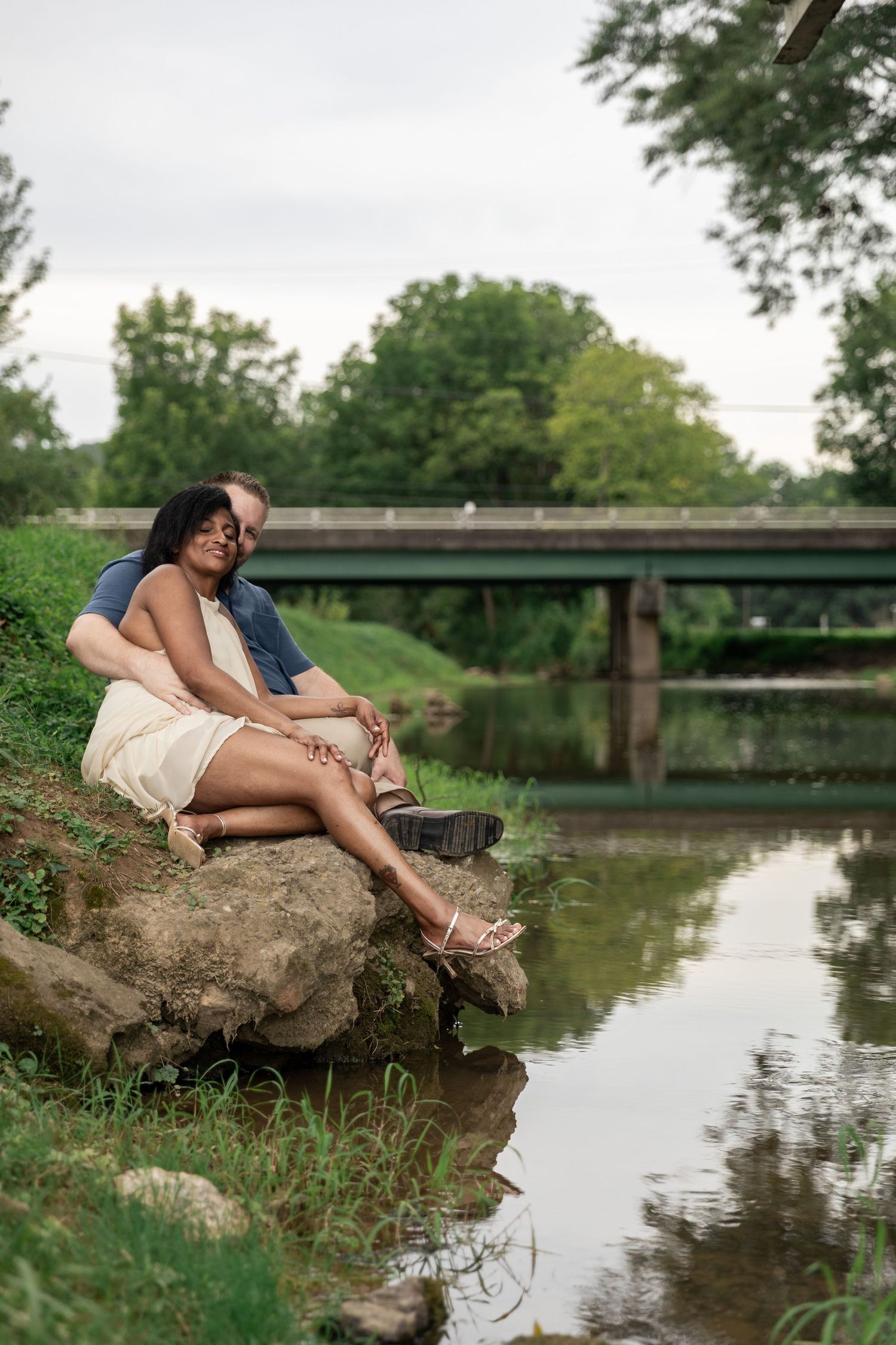 Couple sitting romantically on rocks by a peaceful river, surrounded by lush greenery and a bridge in the background. Rockmart, GA