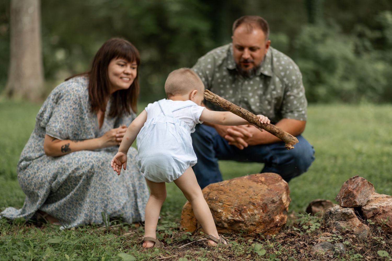 A family with a baby playing outdoors in a park, with parents watching their toddler pick up a stick near rocks.