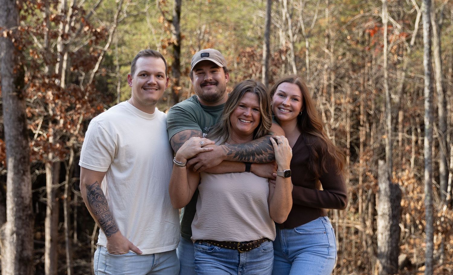 Four smiling adults pose together in an autumn forest with colorful fall foliage in the background.