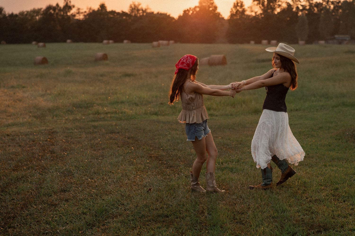 Two women dancing joyfully in a sunlit field at golden hour, wearing cowboy hats and boots on a farm.
