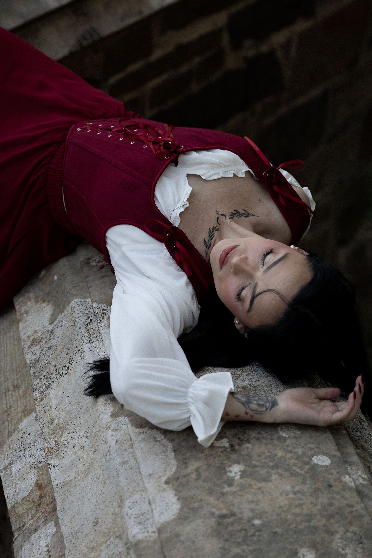 Woman in red and white medieval costume lying on stone steps with eyes closed and visible tattoos. Berry College Rome, GA