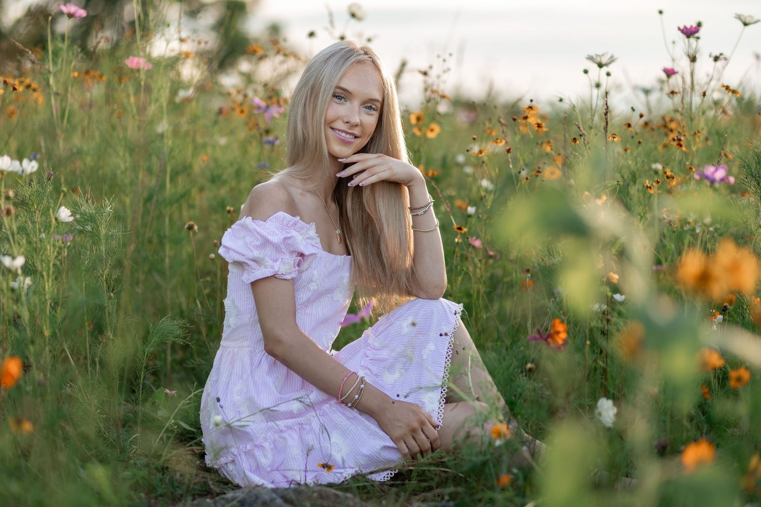Smiling blonde woman in white floral off-shoulder dress sitting in a colorful wildflower meadow on a sunny day.