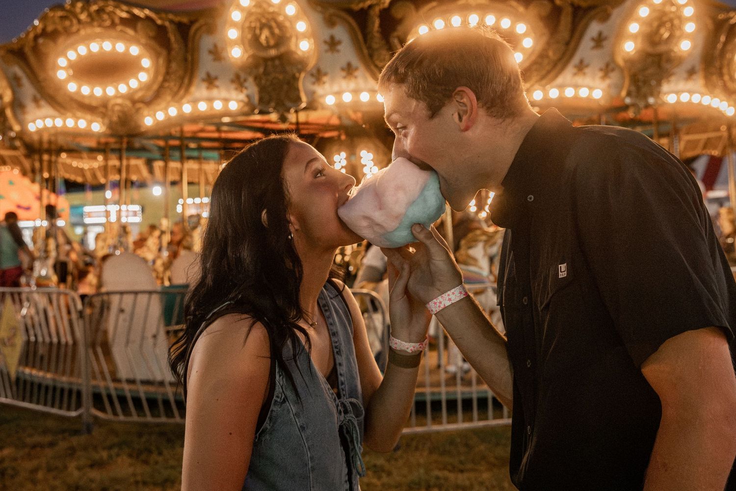 Couple sharing cotton candy at a carnival with a lit carousel in the background at night.