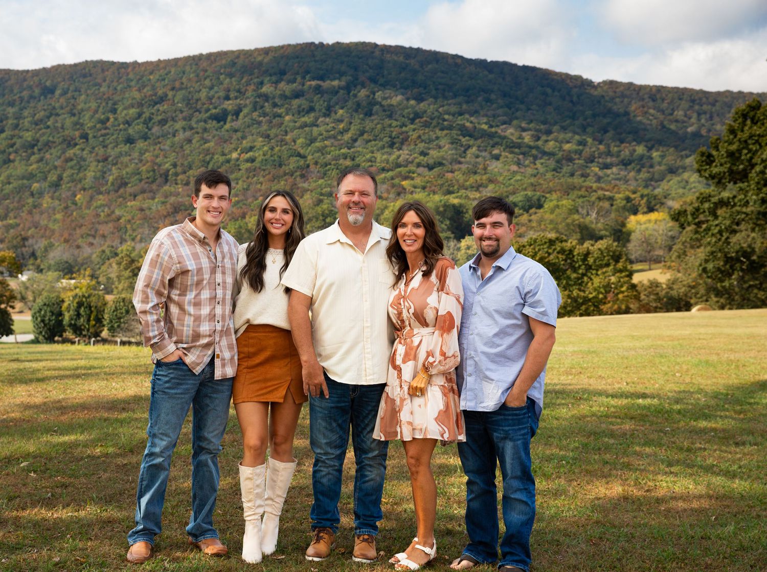 Family of five smiling outdoors on green field with scenic mountain backdrop on a sunny day.