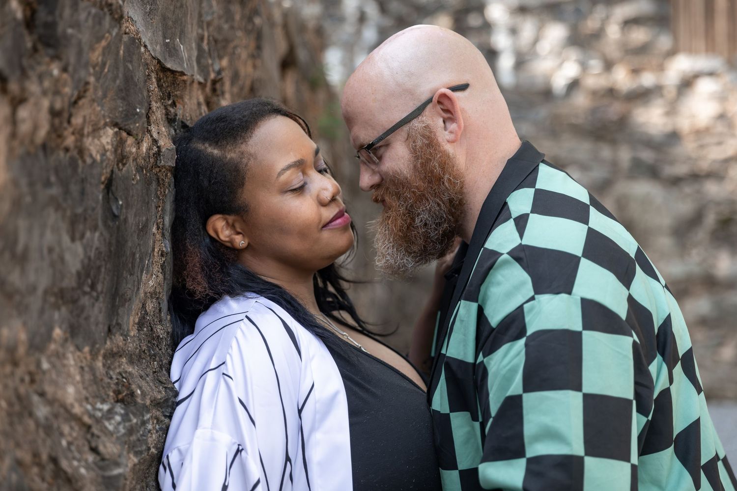 A couple shares an intimate moment, leaning close together against a stone wall outdoors.Euharlee Covered Bridge