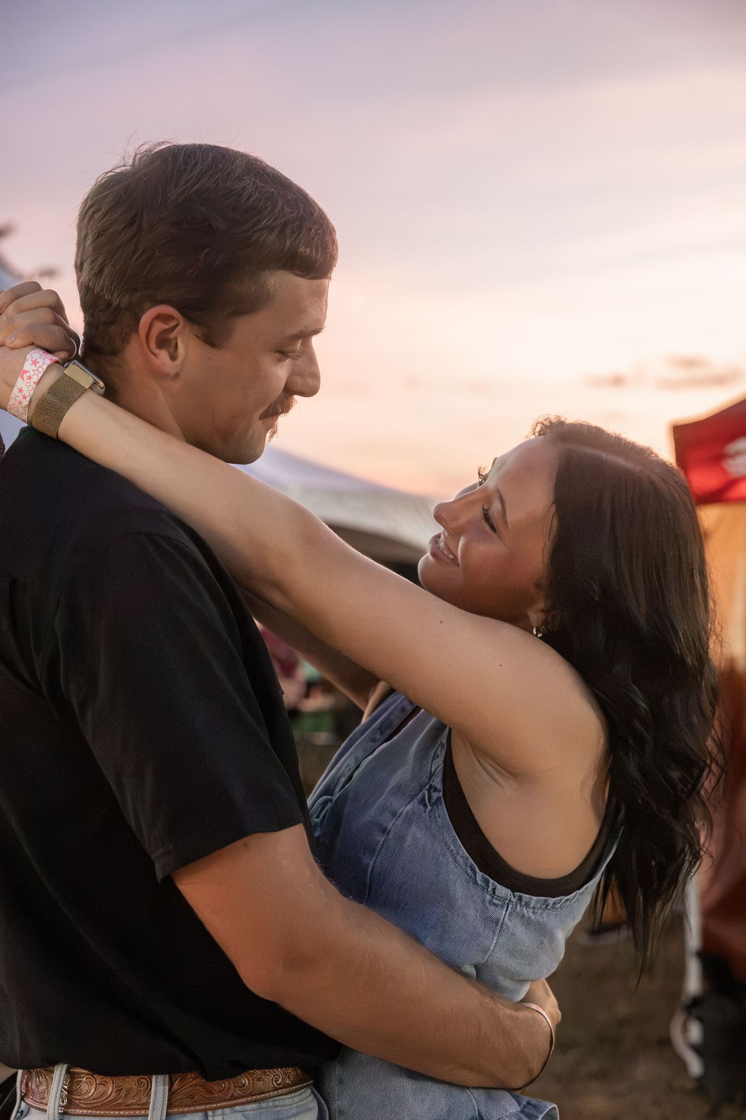 Couple embracing and dancing at an outdoor festival during a beautiful sunset, woman smiling with arms around mans neck.