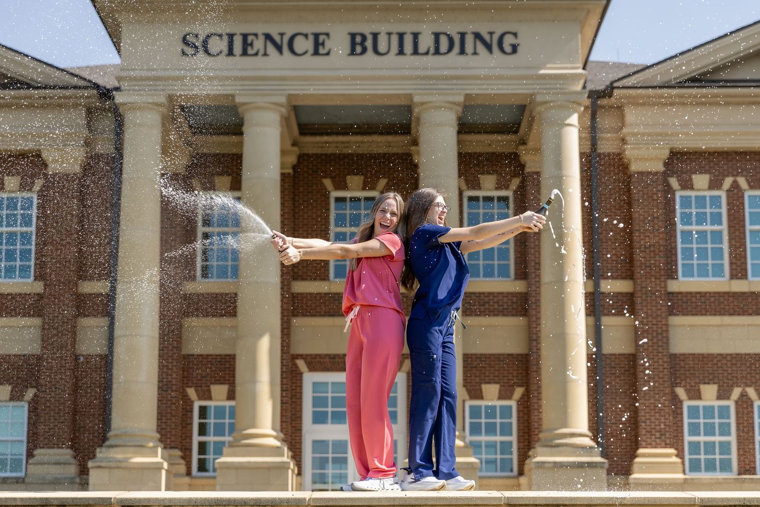 Two graduates celebrate in scrubs popping champagne in front of a brick Science Building on a sunny day- College Grads Northwest Georgia