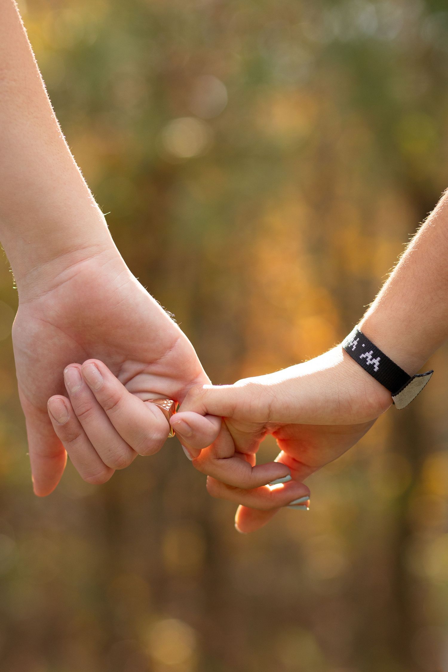 Two people holding pinkies together outdoors in autumn, one wearing a black fitness tracker bracelet.