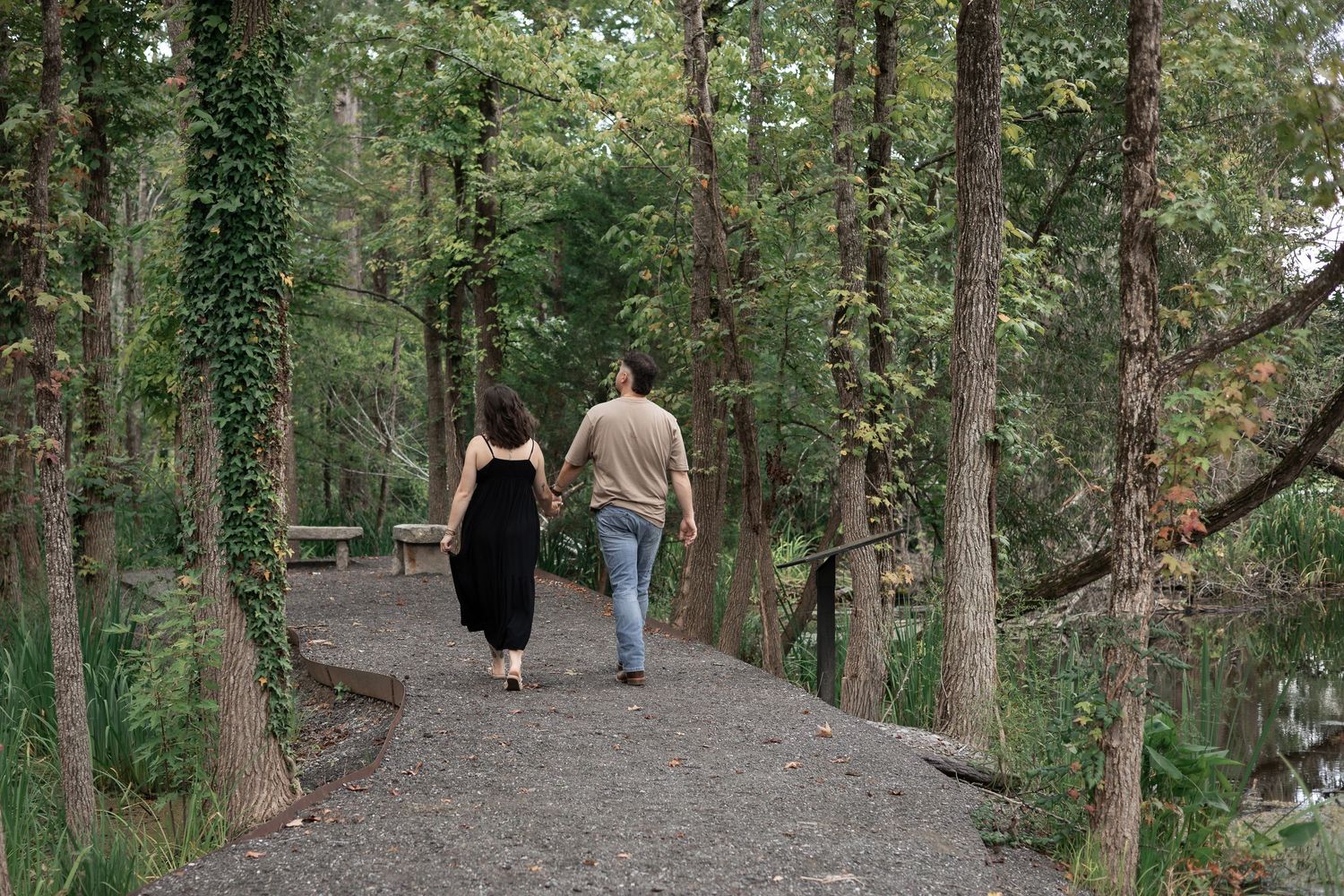 Couple walking hand in hand on a gravel forest path surrounded by lush green ivy-covered trees.