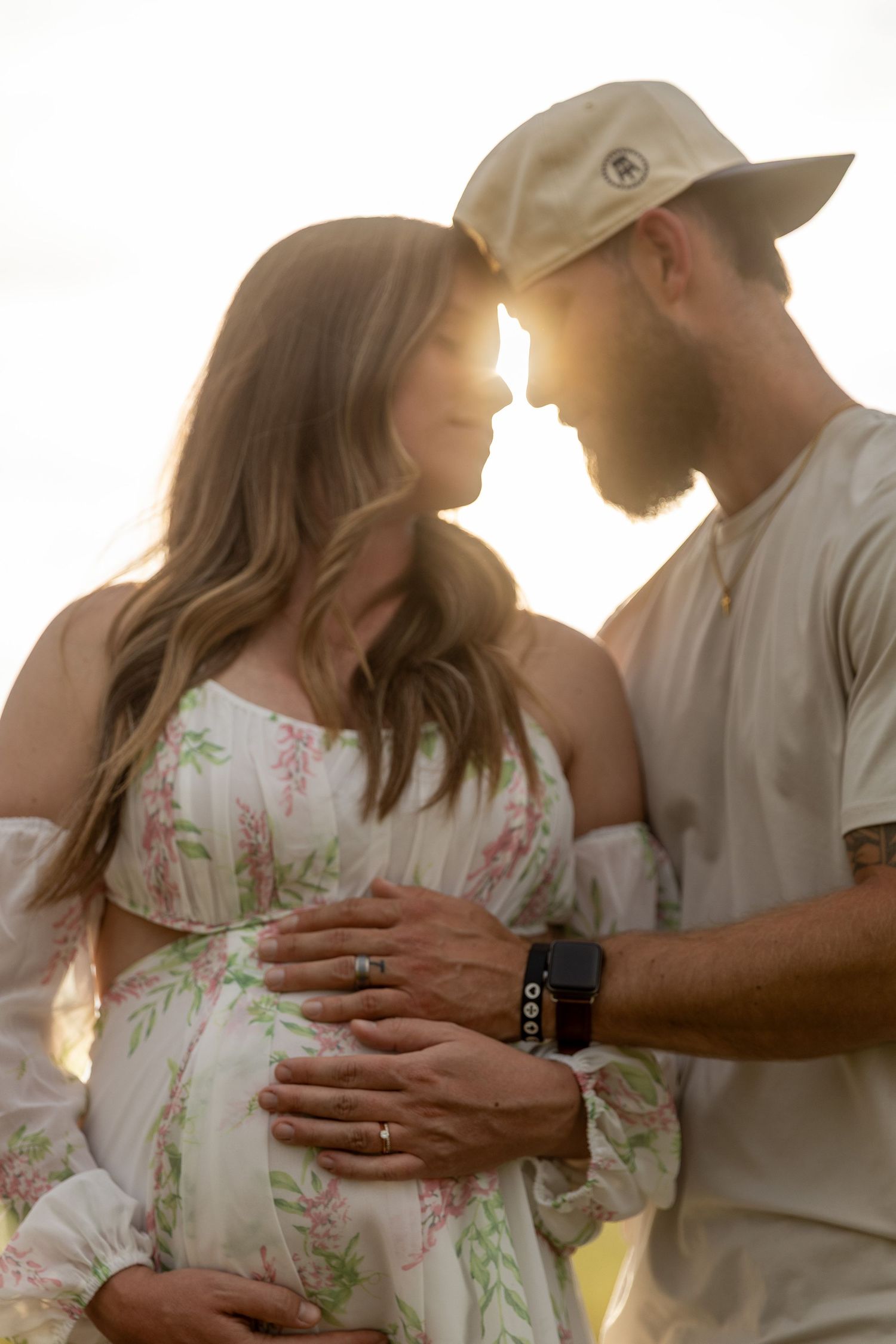 Pregnant couple tenderly touching foreheads at golden hour, hands on baby bump, backlit by warm sunlight.