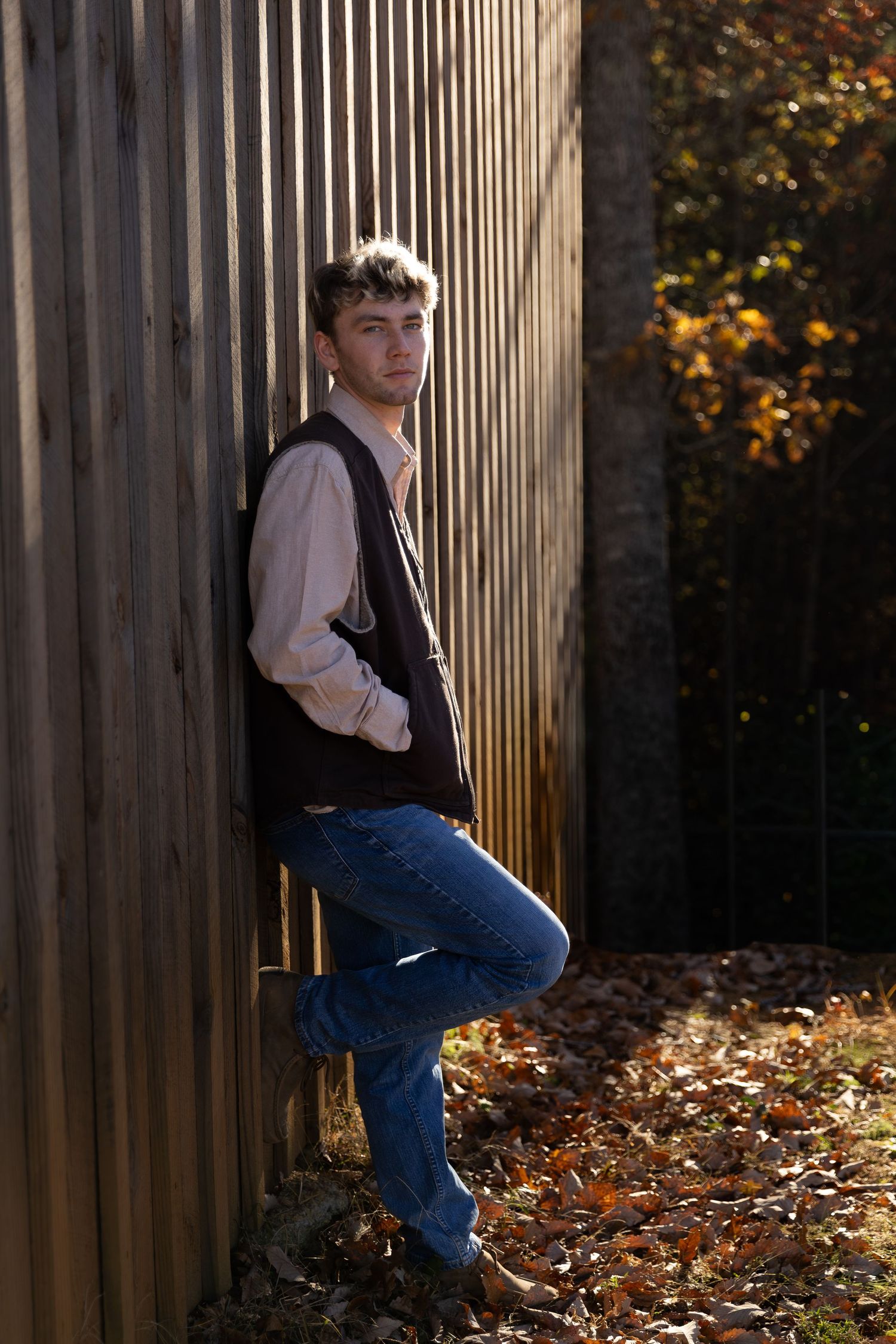 Young man in jeans and vest leaning against wooden fence surrounded by autumn leaves- senior portraits, Northwest Georgia