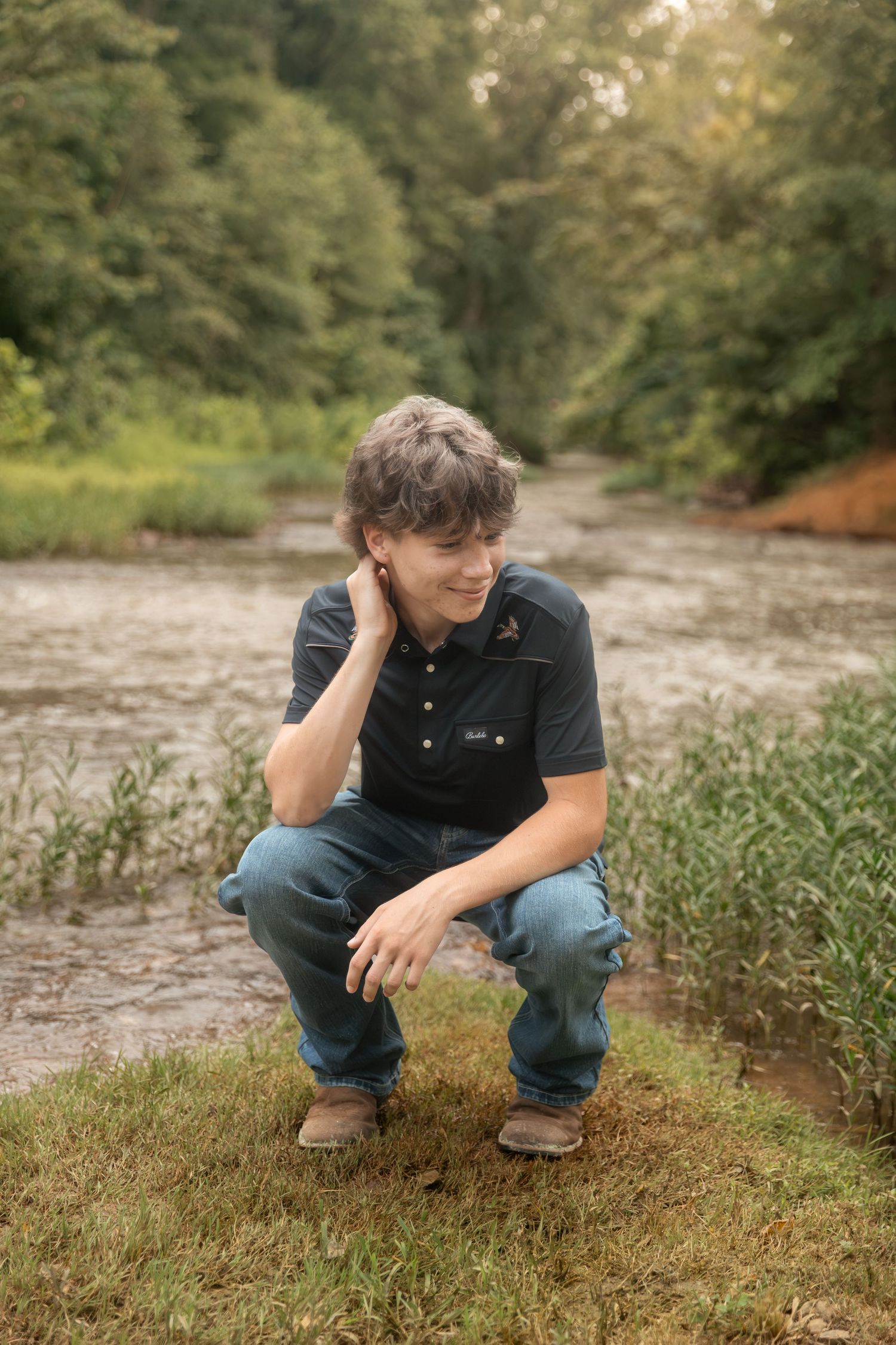 A young man crouching by a creek in jeans and dark polo shirt, surrounded by lush green trees and grass.