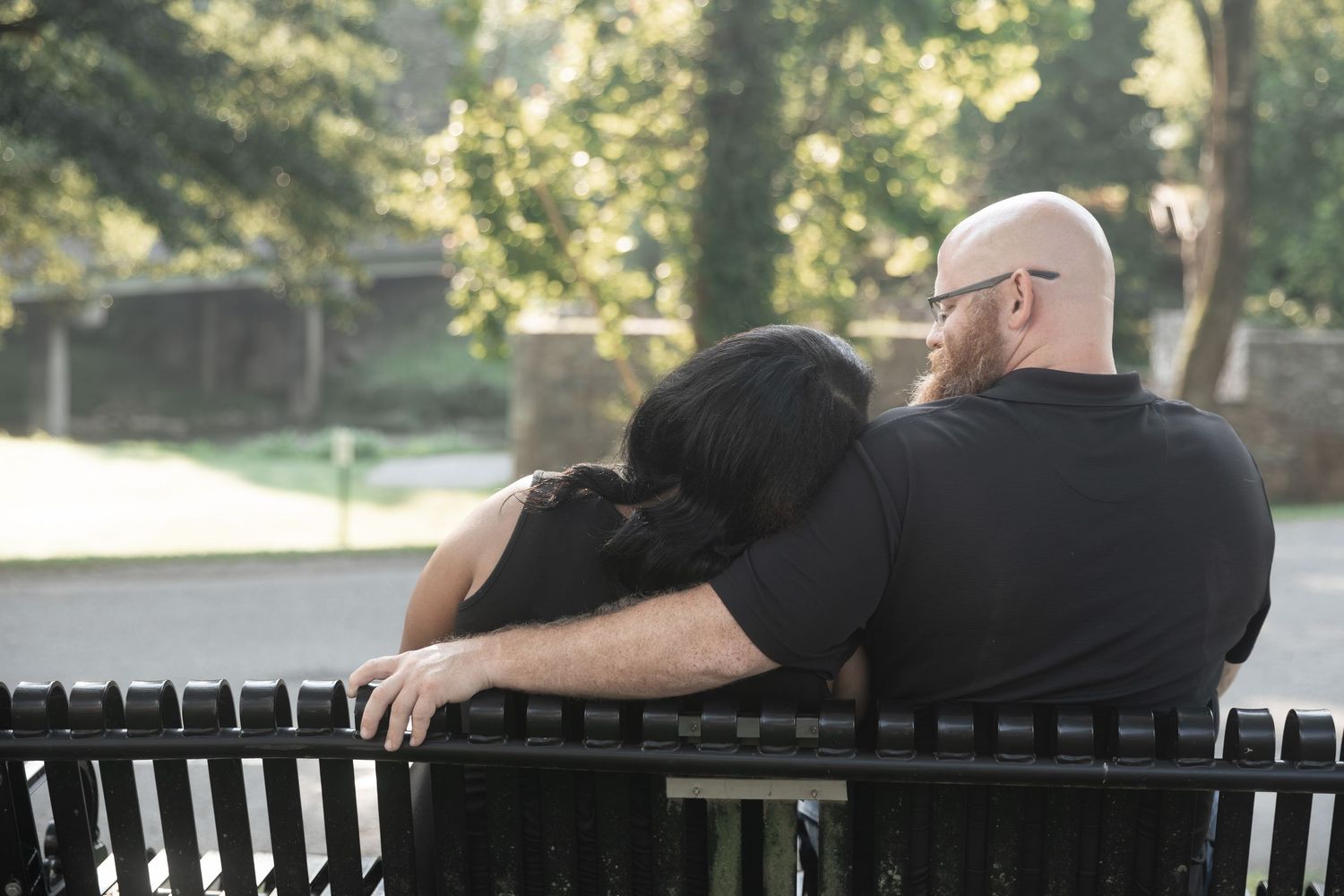 Couple sitting on a park bench, woman resting head on bald man's shoulder, surrounded by lush green trees. Euharlee Covered Bridge