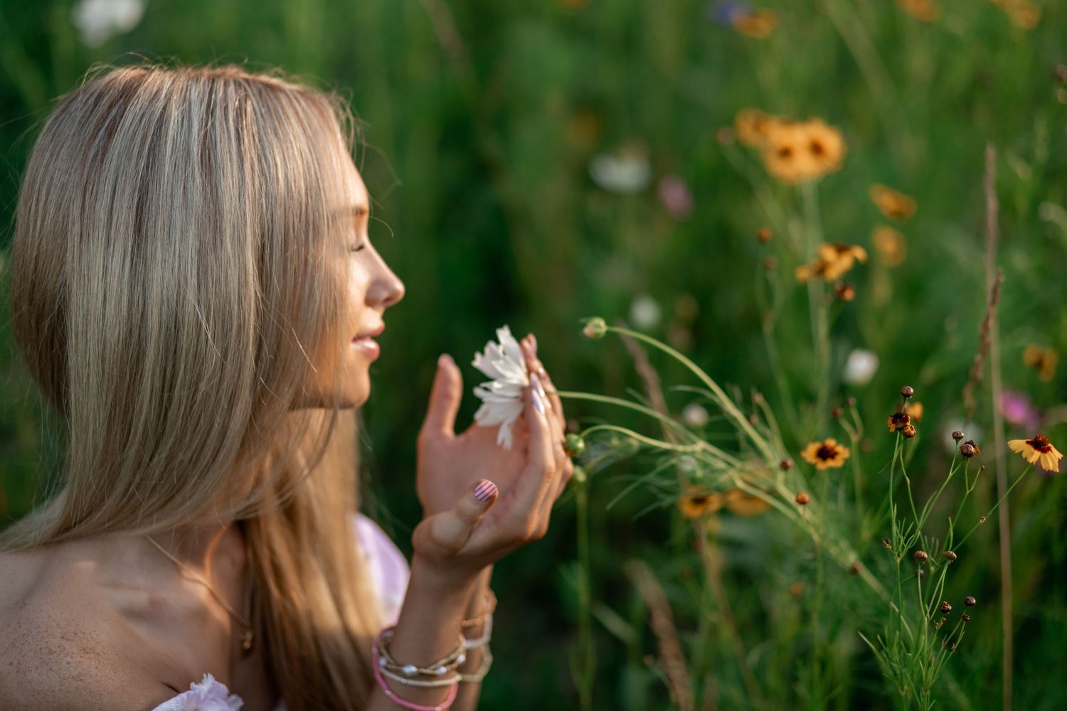 Young blonde woman smelling a white daisy in a vibrant wildflower meadow on a sunny day.