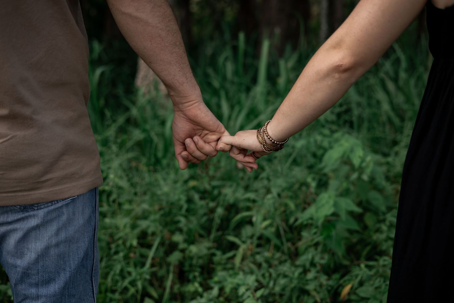 A couple holds hands tenderly in a lush green forest setting, showcasing a romantic outdoor moment.