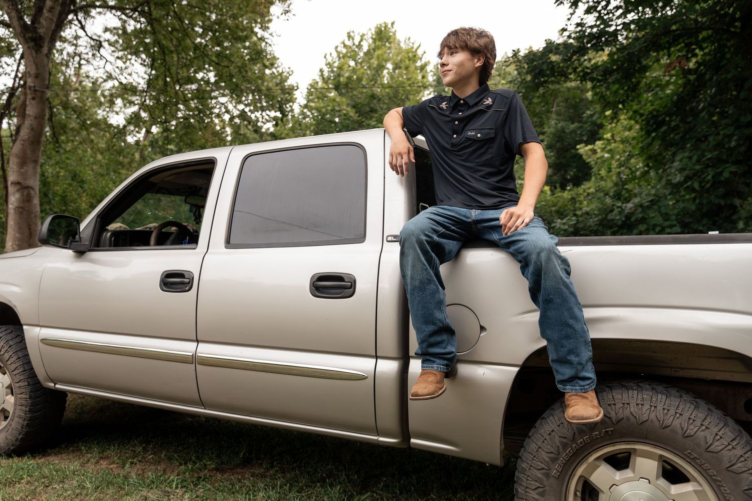 Young man in jeans and polo shirt sitting on the hood of a silver GMC pickup truck outdoors.