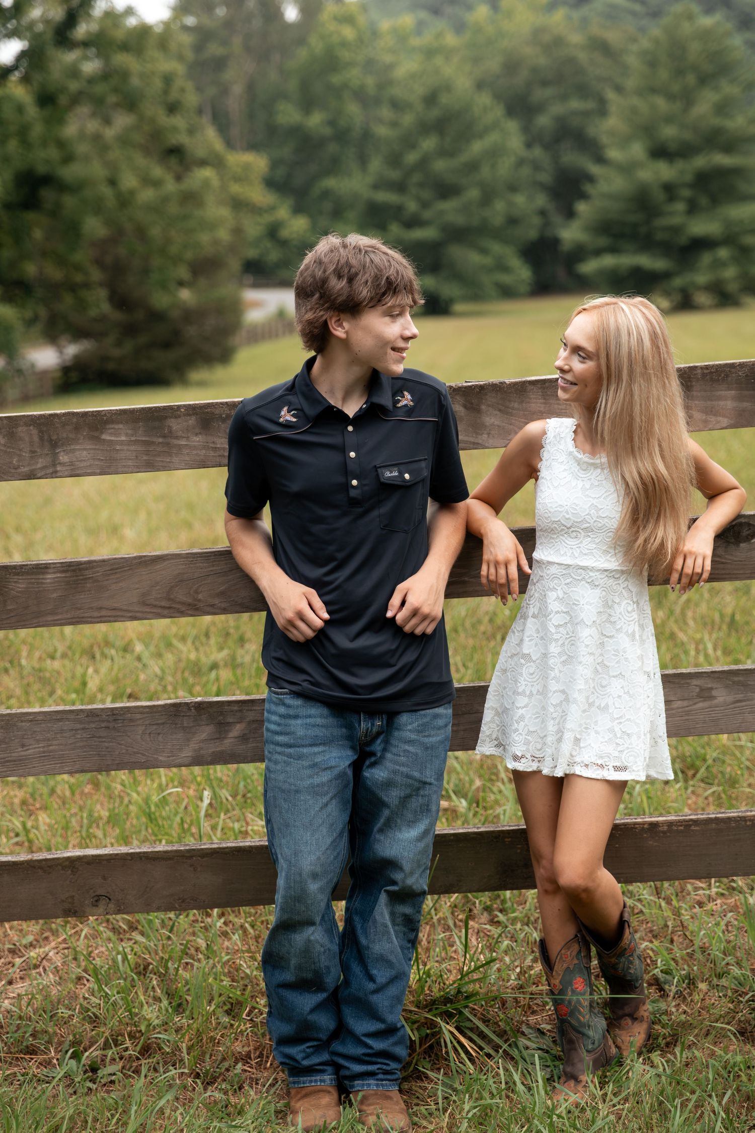 A teenage boy and girl smile at each other leaning on a rustic wooden fence in a green rural countryside setting. Rome, GA