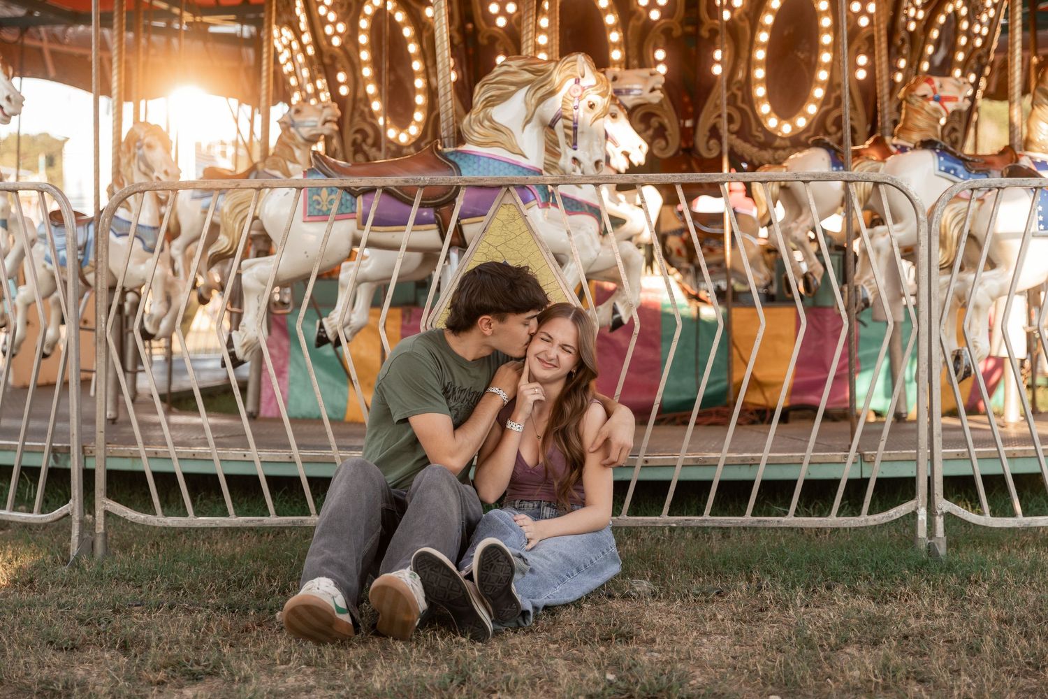 Couple sitting on grass in front of a colorful carousel at a sunny fairground, sharing a romantic moment.