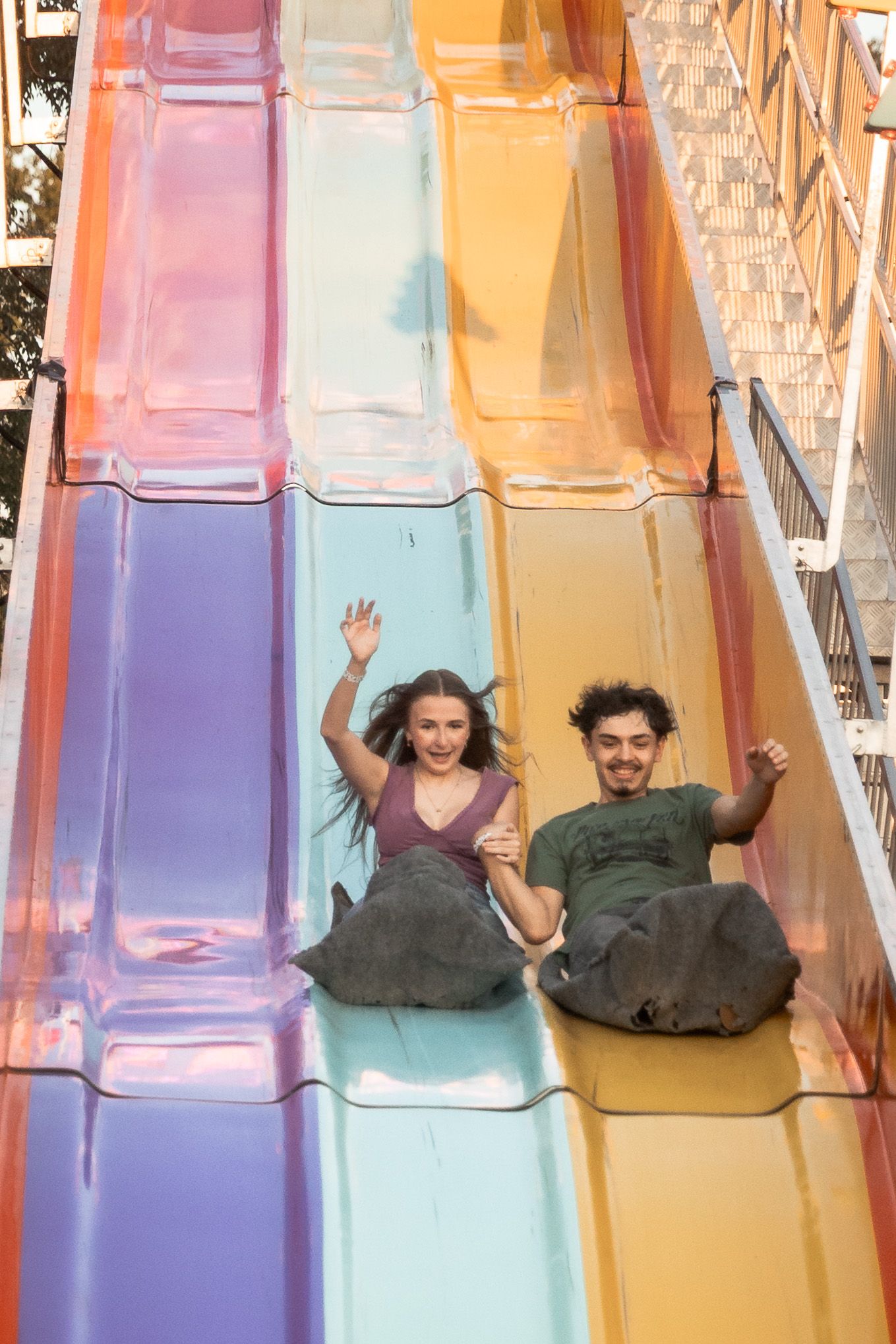Two young adults laughing and waving while sliding down a colorful rainbow slide at an amusement park.