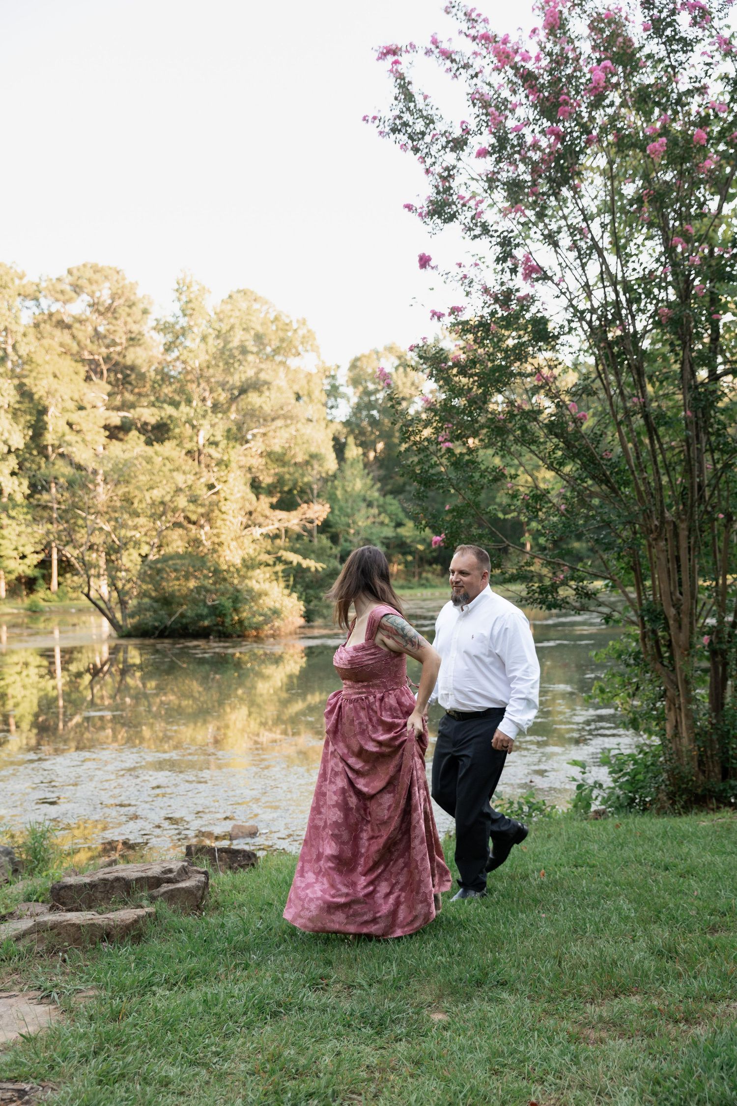 Couple walks hand-in-hand by a serene pond, woman in pink gown, man in white shirt, surrounded by lush trees. Berry College, Rome, GA