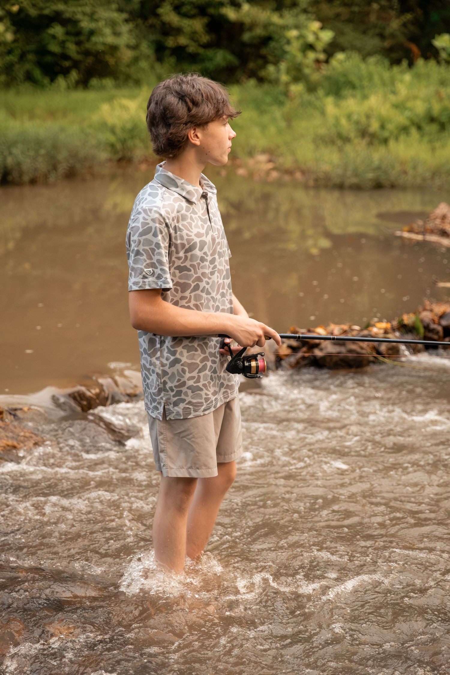 Teen boy wading in a shallow creek while fishing with a spinning rod, surrounded by lush green trees.
